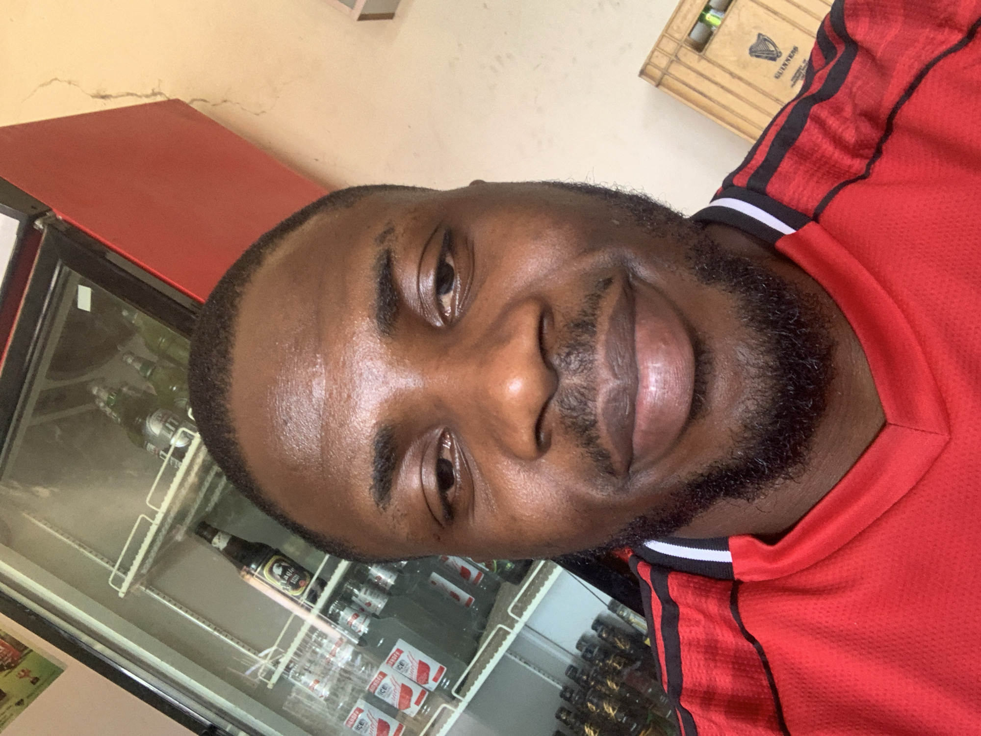 A close-up selfie of a Black man with short, dark hair and a beard, wearing a red shirt with black and white trim. He is smiling slightly. Behind him is a refrigerated display case filled with drinks, including bottles and cans. There's also a wooden crate visible in the background. The location is likely a store or bar, in New Stadium, Nigeria.