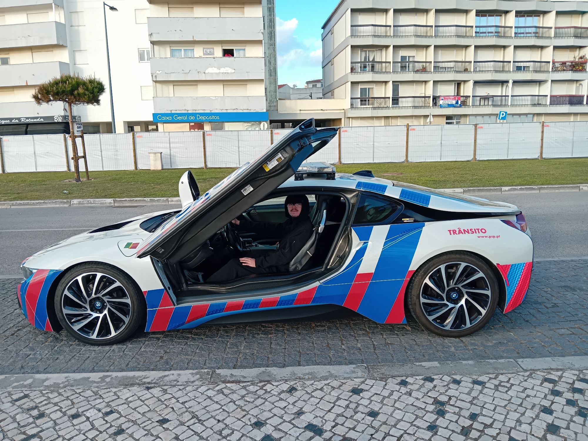 The image shows a white BMW i8 police car, with its gull-wing doors open, parked on a cobblestone street. The car is adorned with blue and red stripes, along with the word "TRÂNSITO" (traffic) and the website "www.psp.pt" on its rear. A person wearing black attire and glasses is seated in the driver's seat.
In the background, there are buildings, including one with a blue sign for "Caixa Geral de Depositos" (a bank). A construction fence is visible in front of a grassy area. A small tree is situated near the sidewalk.
The scene is located in Figueira da Foz, Portugal, indicated by the Portuguese bank and the "TRÂNSITO" labeling of the car.