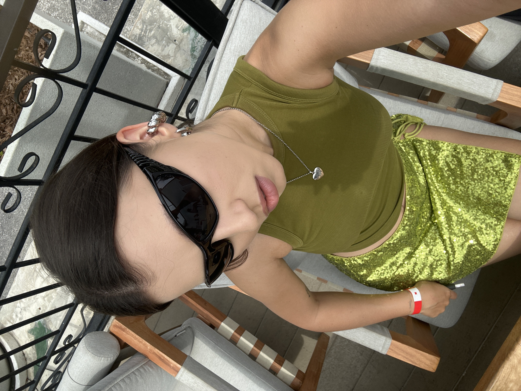 An outdoor, high-angle, medium shot shows a young woman with dark hair pulled back, wearing black wraparound sunglasses, a green sleeveless crop top, and a sequined lime green skirt, sitting on a lounge chair. She is wearing a red and white wristband on her left wrist and a silver heart pendant on a chain around her neck. A large, ornate silver earring is visible on her right ear. The background shows a black metal railing, patio furniture with grey cushions, and a tiled floor. The overall scene suggests a leisurely setting, possibly at a resort or outdoor dining area in Choeng Thale, Thailand.