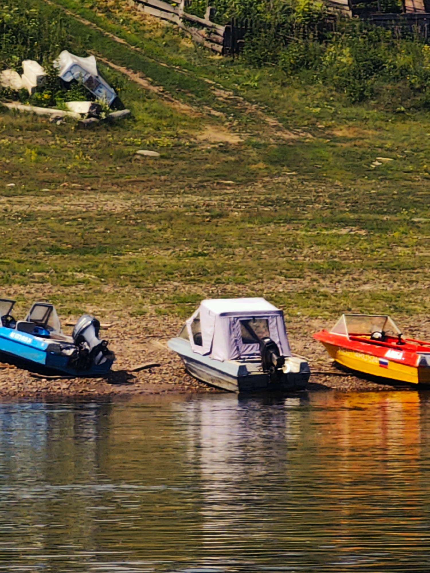 The image depicts a waterside scene in Pogodaevo, Russia, characterized by a calm body of water in the foreground and a grassy, slightly overgrown embankment in the background. Three boats are visible resting on the rocky shore. The leftmost boat is a blue motorboat with a visible outboard motor. Adjacent to it, to the right, is a small boat with a grey canopy. Further to the right, a red and yellow motorboat with a clear windshield is positioned. The embankment behind the boats features sparse vegetation and a wooden fence structure at the top. The water reflects the surrounding environment, creating a textured surface. There are no people visible in the frame.