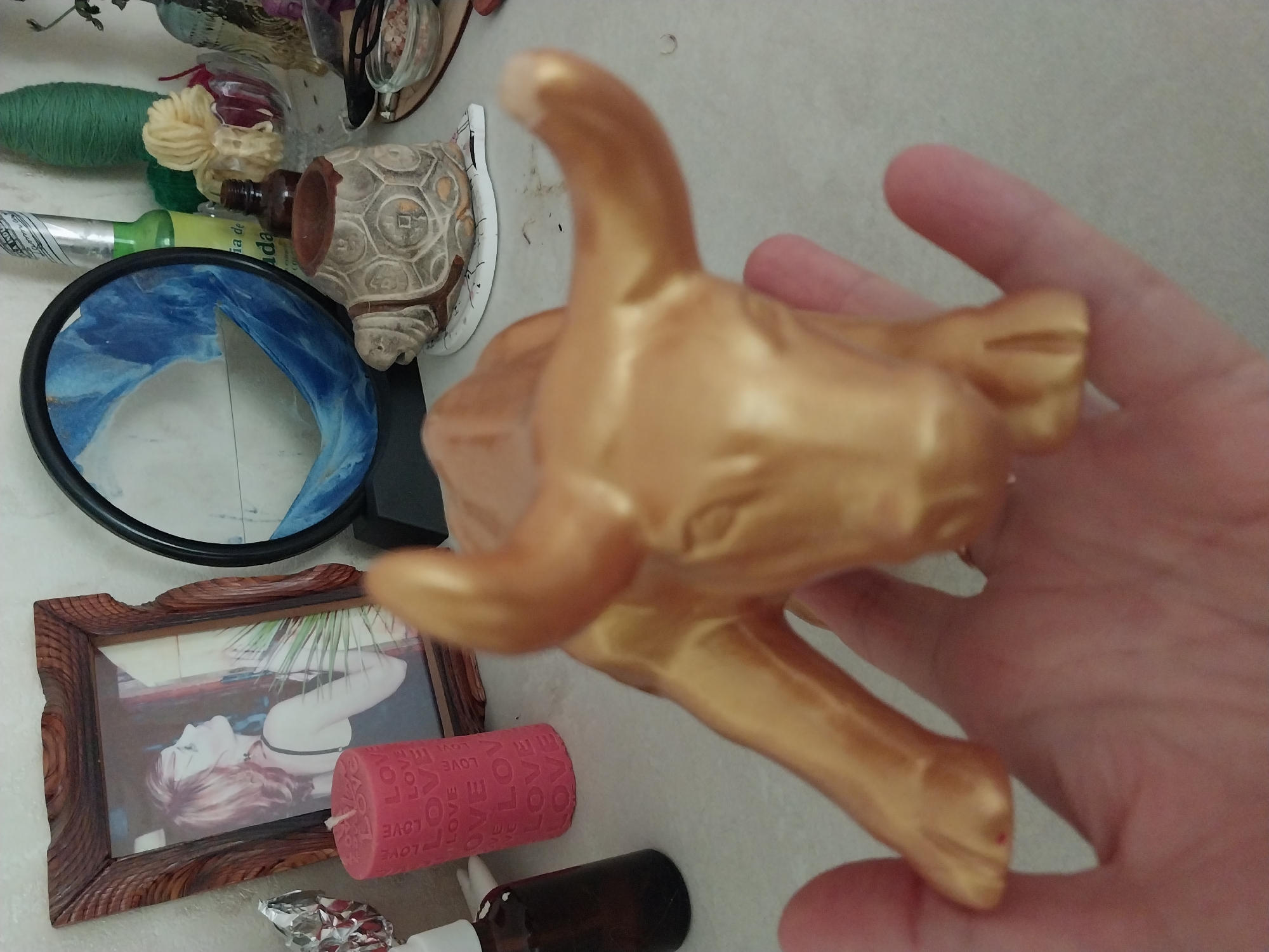 The image shows a close-up of a hand holding a gold-colored bull figurine. The figurine is positioned on a surface that appears to be a tabletop or shelf, with various objects in the background. To the left of the figurine is a pink cylindrical candle with the word "LOVE" repeated on its surface. Behind the candle is a framed photograph of a woman. Further to the right, there is a round mirror with a blue and white abstract design, a turtle-shaped decorative object, and a small glass bowl containing pink crystals. The scene suggests a domestic or personal space, possibly a shelf or dresser. The location in the city of Dnipro, Ukraine is not discernible from the image.