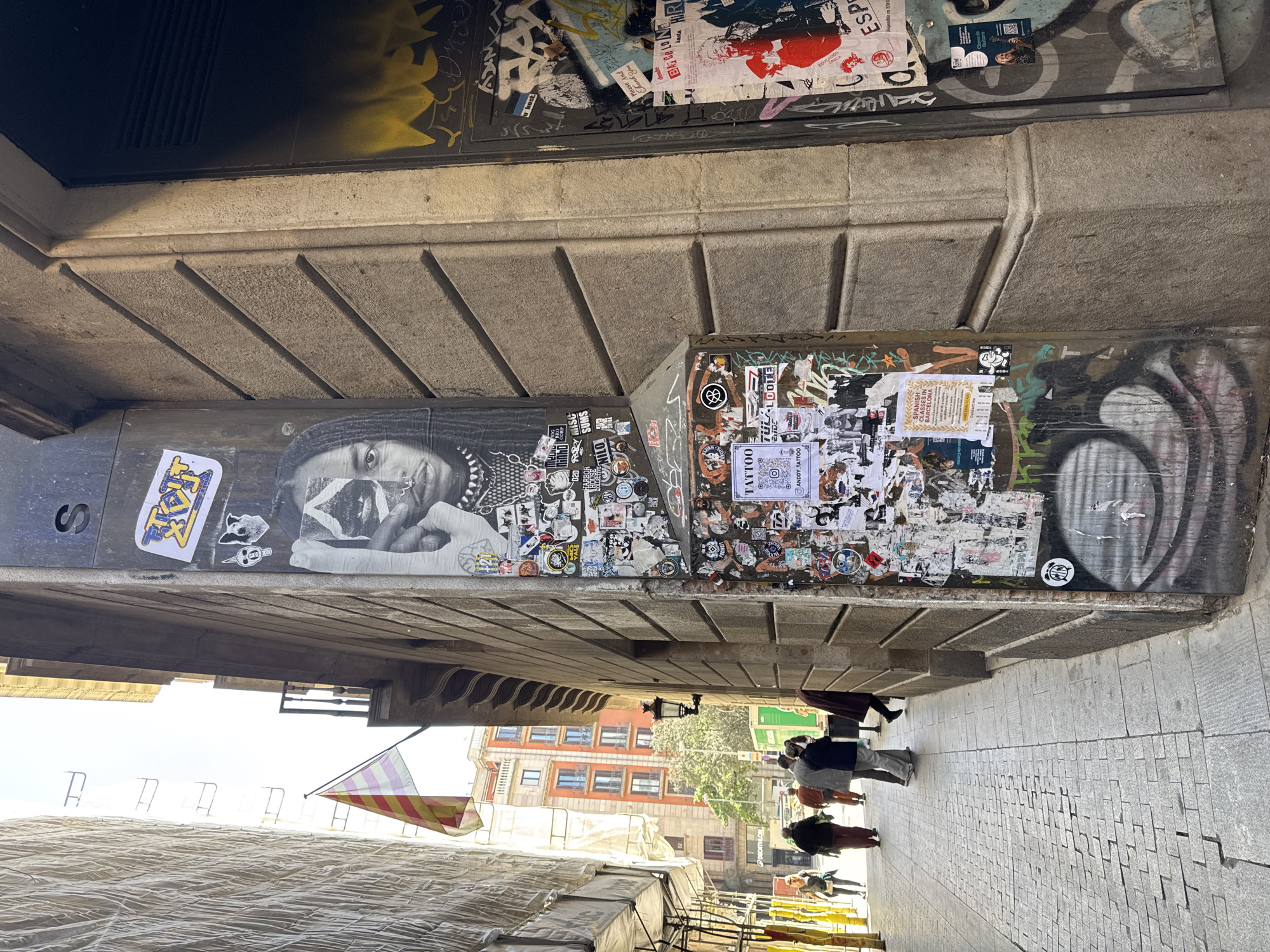 Captured from below an overhanging structure covered in graffiti and stickers in Barcelona, Spain, the image presents a street scene with pedestrians walking on a paved sidewalk. The overhang features art and graffiti, with stickers densely packed on its surface. From left to right there is what appears to be a black and white stencil of a person wearing a mask and bandanna, with the word TAXI on a yellow and blue background. Continuing further right is a patch of dense graffiti and stickers, and then a graffiti design in the shape of a ring. In the background, buildings with orange facades are visible, and a Catalan flag is flown. A large, draped structure or tarp obscures part of the foreground. Pedestrians are walking towards the end of the passageway. Signs for the sports chain DECATHLON are visible in the distance.