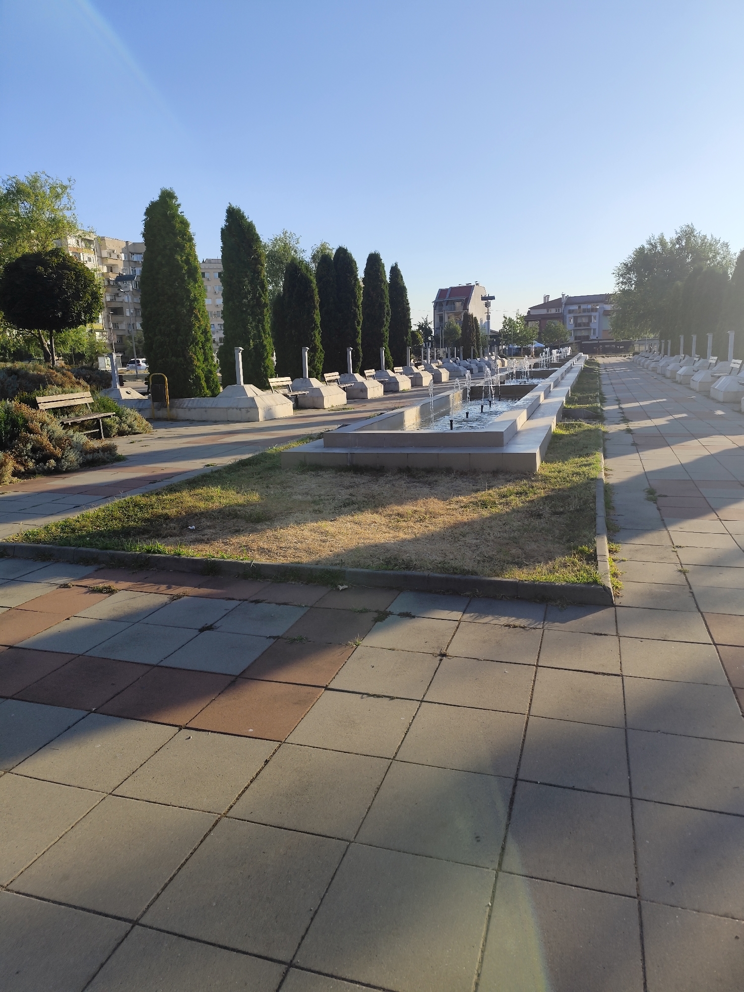 The image depicts a sunny day in a city park or public square in Montana, Bulgaria. The foreground is dominated by a paved walkway with alternating light gray and reddish-brown square tiles, casting long shadows from the early morning or late afternoon sun. To the left, a section of dry grass is bordered by a concrete curb. Beyond the grass, several park benches are visible, some with people sitting on them. A line of tall, slender evergreen trees runs along the left side of the walkway, interspersed with white, monumental structures. In the middle ground, a long, rectangular fountain features multiple jets of water spraying upwards, with more white structures and benches lining the right side of the fountain. Further in the background, apartment buildings and other urban structures are visible under a clear blue sky with a faint rainbow arc. The scene suggests a well-maintained public space designed for relaxation and recreation.
