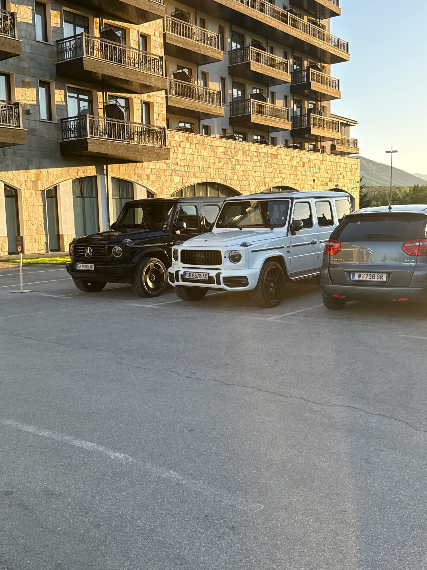 The image depicts an outdoor parking area in front of a multi-story building constructed with stone and concrete. The building features multiple balconies with railings. The scene is illuminated by natural sunlight, casting long shadows. In the foreground, three vehicles are parked: a black Mercedes-Benz G-Class, a white Mercedes-Benz G-Class, and a gray Citroën C4 Picasso. The license plates of the vehicles are visible. The parking lot surface is asphalt with painted white lines. In the background, trees and mountains are visible, suggesting a natural or semi-rural environment. The image was taken in Razliv, Bulgaria, as indicated by the context. There are no people visible in the frame.