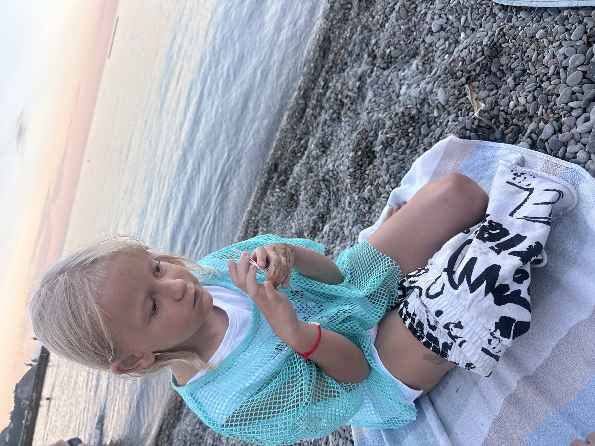 The image shows a young girl with blonde hair sitting on a pebble beach in Partenit, Ukraine. She is wearing a white t-shirt, a sheer turquoise cover-up, and white shorts with a black graphic print. The girl is holding a small object in her hands and looking down at it. She has a red bracelet on her left wrist and what appears to be henna on her right hand. The setting is a beach with the sea in the background. The water is calm and reflects the soft light of what appears to be either early morning or late evening, with a pastel sky suggesting a sunrise or sunset. The overall mood is peaceful and contemplative. There is no visible text in the image.