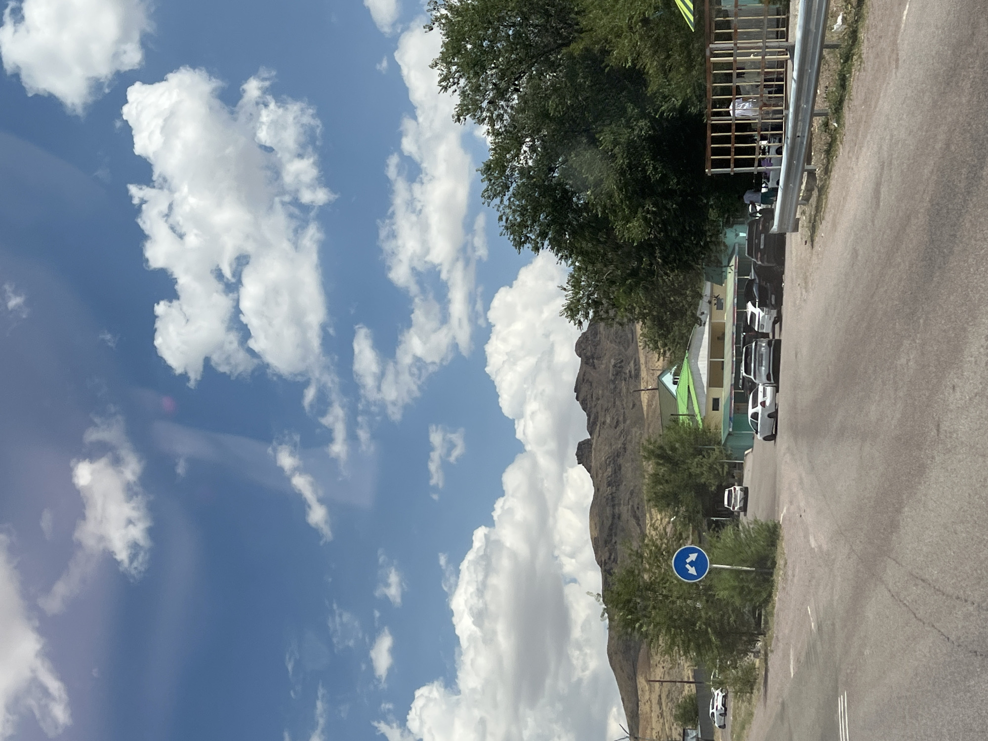 The image depicts a roadside scene in Karlygash, Kazakhstan. The sky is blue with scattered white clouds. In the foreground, a paved road curves to the right. To the left of the road, a round blue traffic sign with white arrows pointing left and right is visible, surrounded by green foliage. Further ahead, several cars are parked or driving on the road, including white sedans and darker SUVs. Beyond the road, a few low-lying buildings with green and light-colored roofs are situated. A line of trees runs along the middle ground, separating the road area from a rocky, mountainous landscape in the background. To the far right of the image, a metal fence encloses an area with some individuals standing near what appears to be a shaded structure.