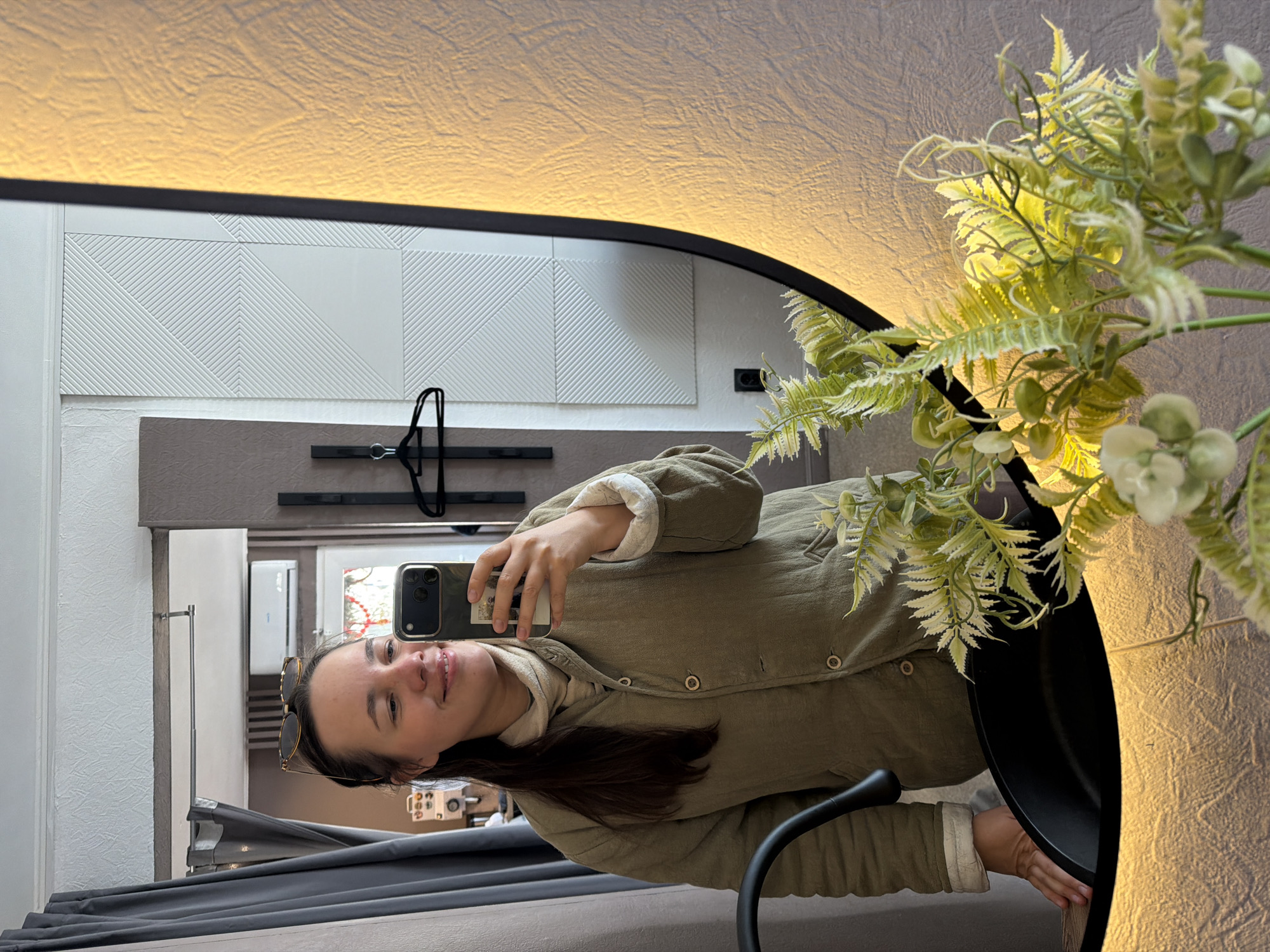 A young woman is reflected in a mirror, taking a selfie with her smartphone. She is wearing a khaki green quilted jacket with light-colored lining visible at the collar and cuffs. The woman has brown hair and is smiling slightly. Behind her in the reflection is a wall with a geometric pattern in white tiles, below which is a brown-toned panel. A black clothes hanger is visible. The environment includes a plant in a black pot positioned beside the mirror, partially obscuring the view. The location of the scene is possibly in Chaikovsky, Russia, although this cannot be definitively determined from the image alone. The mirror has a black frame with a warm-toned light.