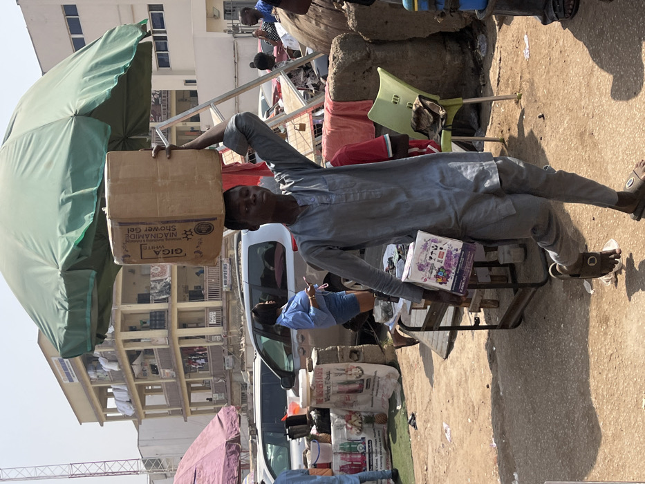 An outdoor market scene is depicted under a clear, bright sky in Abuja, Nigeria. The primary subject is a young male, likely in his late teens or early twenties, with dark skin, wearing a grey traditional tunic and matching trousers, along with dark slip-on sandals. He is walking towards the left of the frame. On his head, he balances a large, rectangular brown cardboard box, secured by his raised left hand. The box has a label partially visible that reads "GIGA WHITE NIACINAMIDE Shower Gel." In his right hand, he carries a smaller, rectangular purple and white box, with "FLEXOM LTD" visible on its side.
In the midground, to the left of the main subject, a market stall with a white banner displays "THE MASTER'S TASTE" alongside images of pineapples and various bottled beverages. Colorful plastic bottles are arranged on the stall. A silver SUV or van is parked behind this stall. To the right, a woman with short dark hair, dressed in a light blue short-sleeved top and denim shorts, stands near parked vehicles, looking at a smartphone. Further right, a light green plastic chair is visible beside concrete blocks and a metal structure draped with red fabric.
The background features a three-story, light-beige building with numerous balconies, some adorned with railings, and laundry hanging from various levels. A partial sign on the building's facade appears to read "HUM SUPER STORES." Another building is visible to the right. A large green market umbrella partially shades the upper central portion of the scene, with a reddish-purple umbrella visible on the far left. The ground is an uneven, dusty, light-brown surface, indicating a mixture of dirt and worn pavement. The strong shadows suggest a high sun angle. The overall environment indicates a busy commercial street or market area within Abuja.