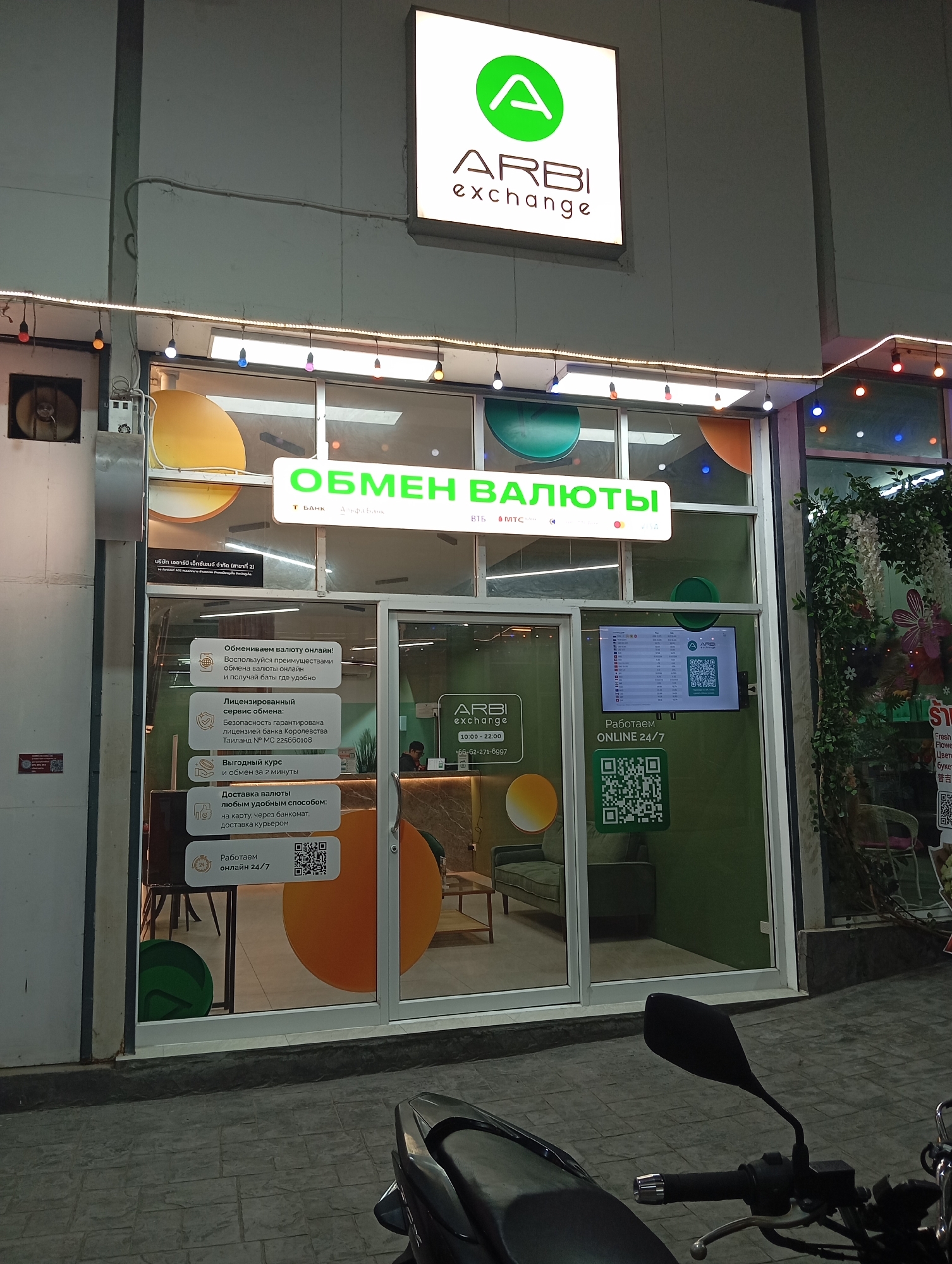 This is a nighttime street-level exterior shot of an Arbi Exchange currency exchange office located in Karon, Thailand. The building features a prominent illuminated sign with the Arbi Exchange logo and name. Below the sign, a large glass storefront displays various informational posters and a digital screen showing currency exchange rates and QR codes. The office appears to be open, with a person visible inside. The street in front of the office is paved with interlocking stone tiles, and a motorcycle is parked in the foreground, obscuring the lower part of the storefront. The facade of the building is decorated with string lights.
