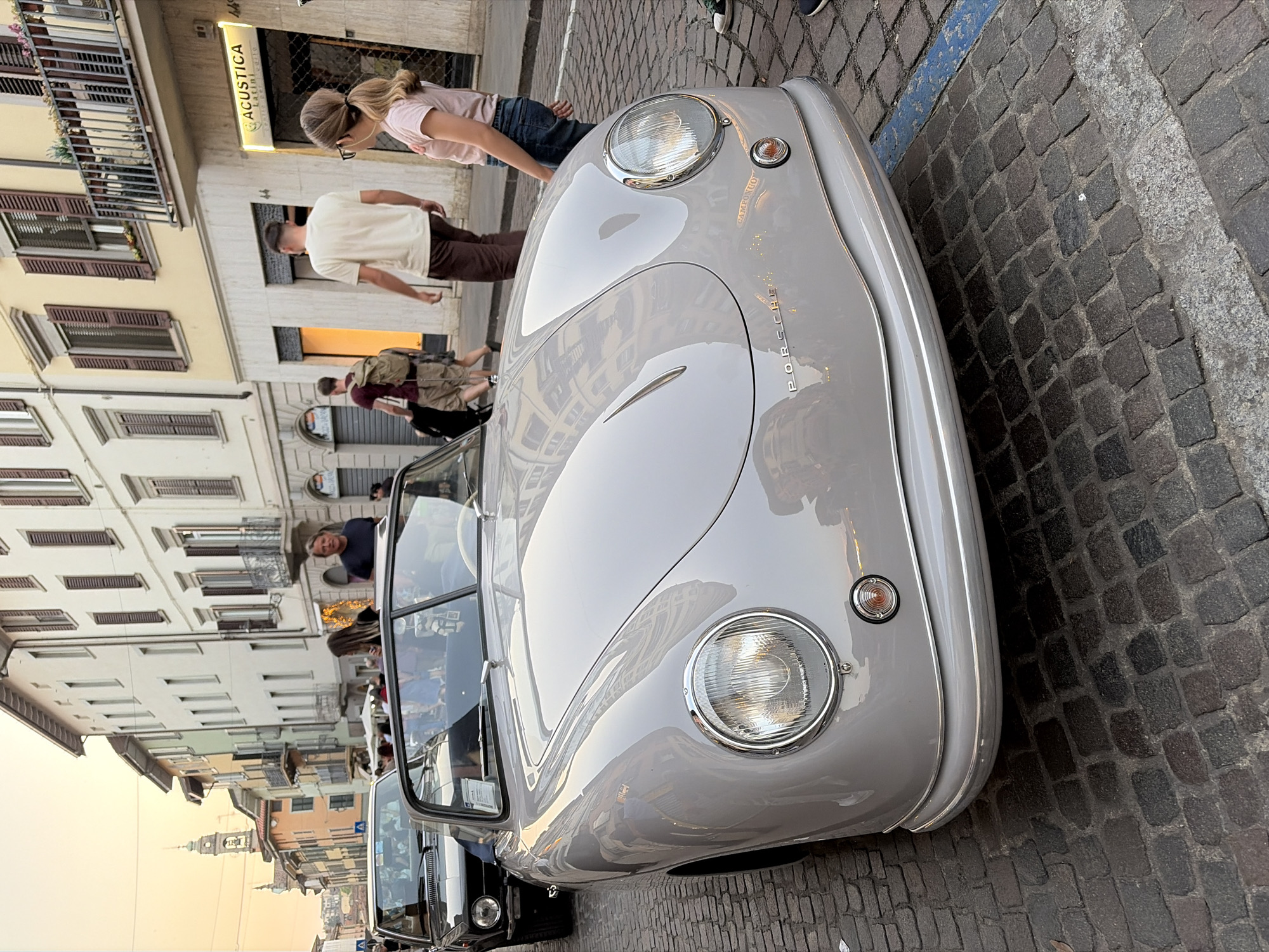 The image depicts a gray Porsche 356 parked on a cobblestone street, likely in Bergamo, Italy, based on the architecture visible in the background. The car is parked at an angle, with its front facing the upper right corner of the frame. In the background, several people are walking along the sidewalk, including a blonde woman in a pink shirt and jeans, a man in a white shirt and brown pants, and other individuals walking in groups. Buildings with multiple windows and balconies line the street. A church tower can be seen in the far background. There is a storefront with the word "ACUSTICA" visible.