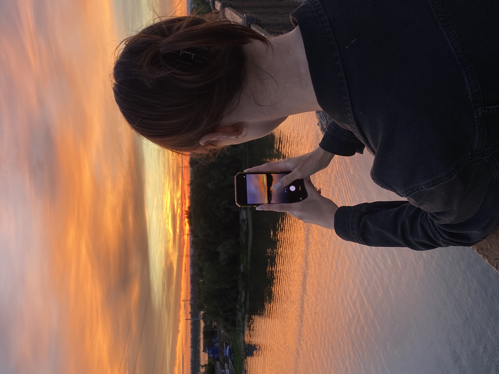A person, viewed from the back, stands on a bridge or elevated walkway overlooking a body of water. The person has dark hair pulled up in a bun with a clip and is wearing a dark denim jacket. They are holding a smartphone in their right hand, capturing a photograph of the sunset. The phone screen displays a vibrant sunset over a reflective body of water. The sky is a vivid gradient of orange and pink, with scattered clouds. In the distance, across the water, is a tree line and some buildings, with power lines visible against the sky. The water itself is a warm orange, reflecting the colors of the sunset. The overall scene is a serene moment of appreciating nature's beauty in the city of Novokuybyshevsk, Russia.