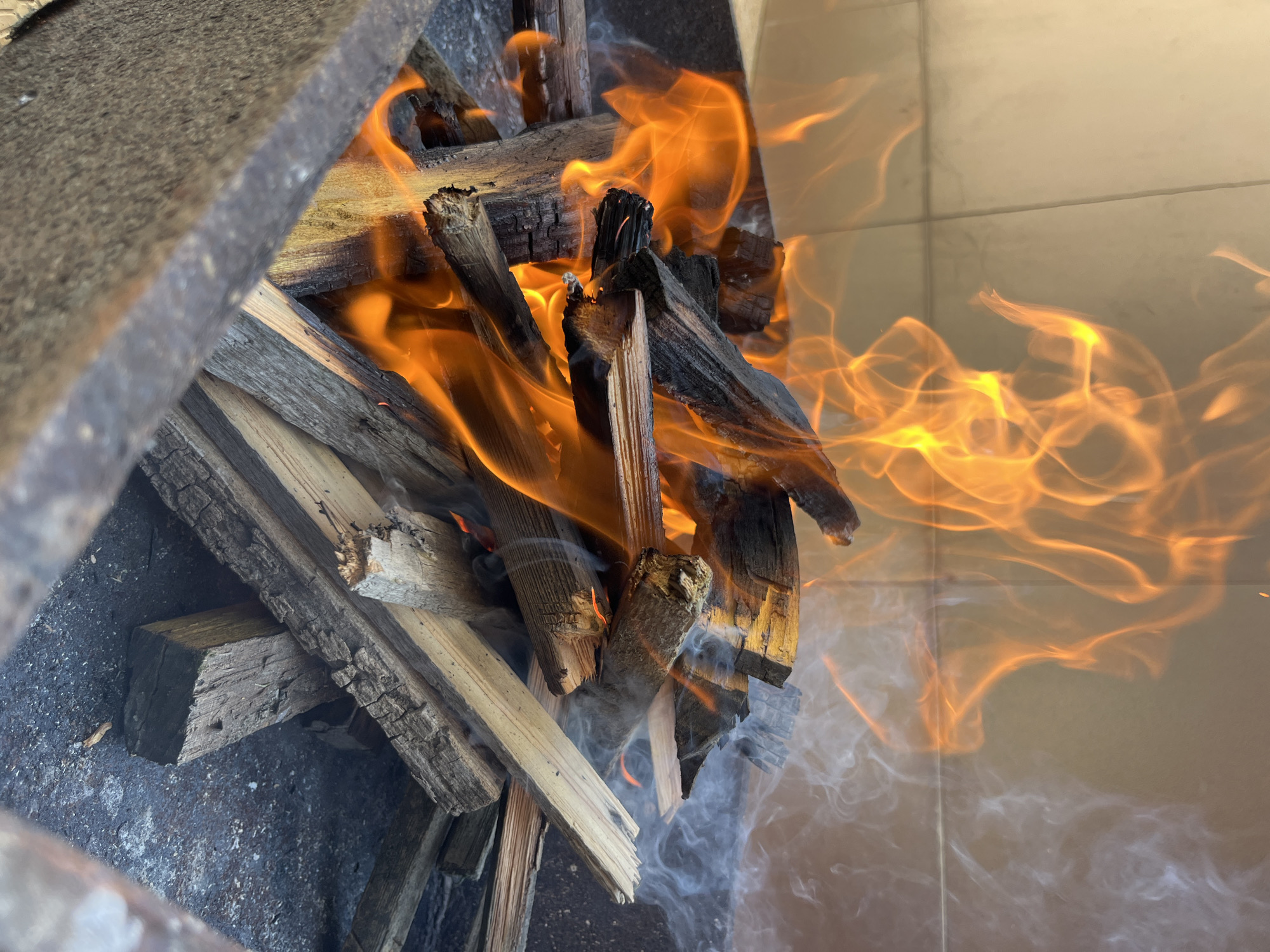 A wood fire is burning inside a fire pit. The wood is arranged in a haphazard pile with flames rising and smoke emanating. The fire pit is constructed of stone. The background is a light-colored tiled surface. The location is Kaspiysk, Russia.