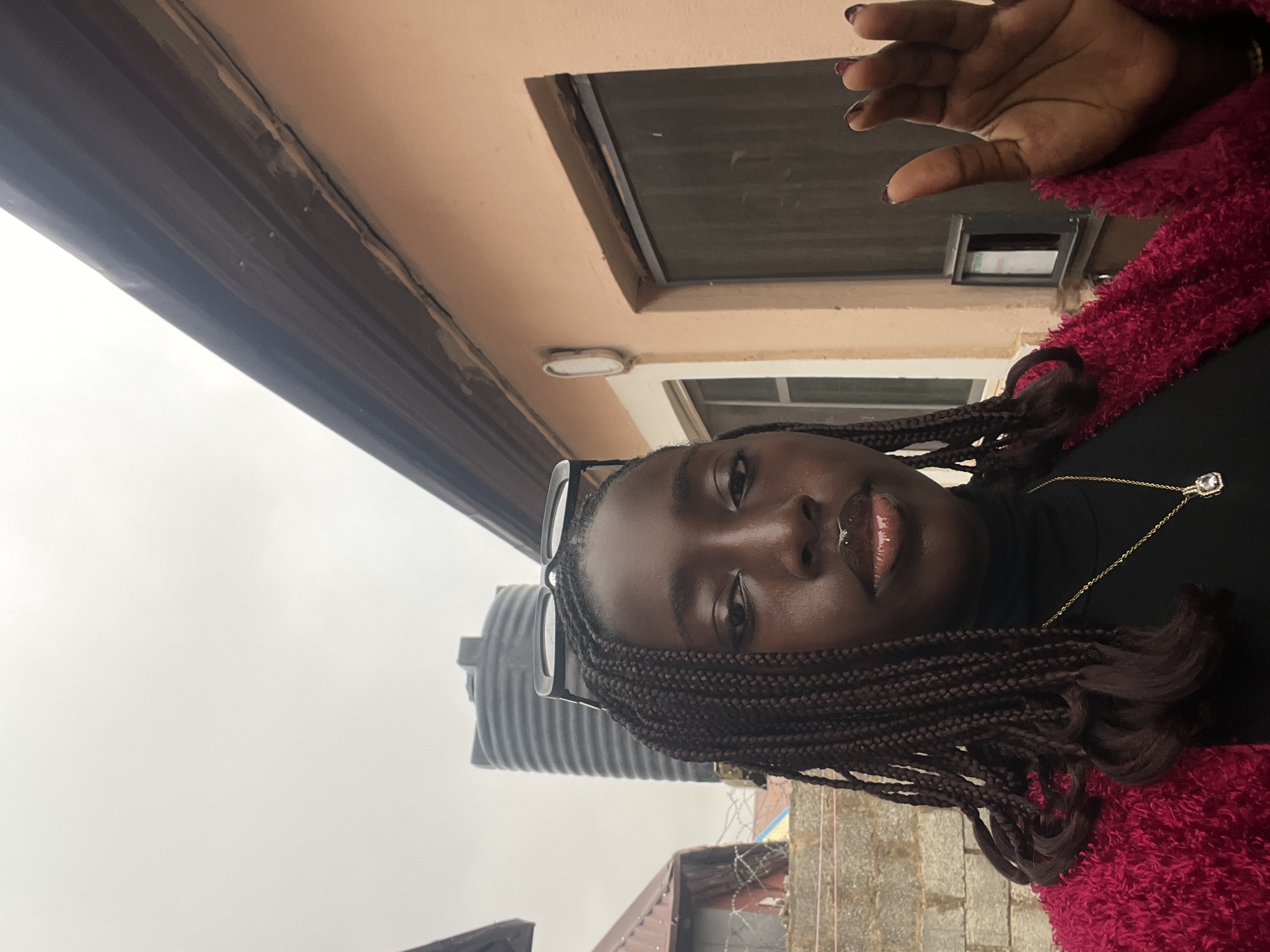 A young woman with dark skin and braided hair is posing for a selfie. She is wearing glasses perched on her head, a black turtleneck, and a gold necklace with a jewel pendant. Her lips are glossy. She is wearing a fuzzy, red jacket. Her hand is raised in the air above her head.
The background includes a peach-colored building with a dark window, a light fixture, and a white window. A large, dark gray water tank is visible behind her. There is also a glimpse of a building with a red roof and barbed wire.
The location is likely in Abuja, Nigeria, based on the style of architecture and the water tank. The sky appears overcast.
