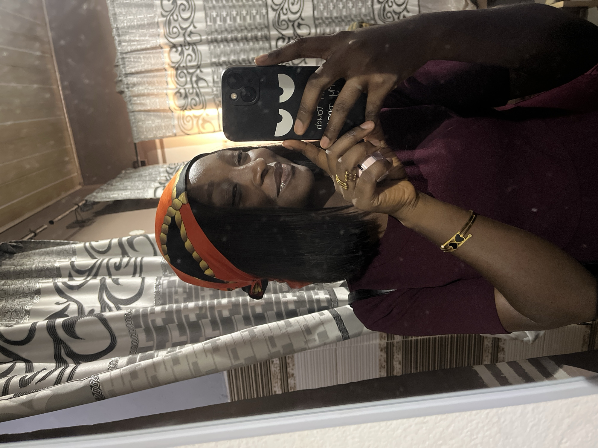 A woman is taking a selfie in what appears to be an indoor setting. She is holding a black smartphone with cartoon eyes on the case. She is wearing a patterned headscarf with orange and black elements, a dark red shirt, and a gold bracelet with black heart accents. She has multiple rings on her fingers. The background includes decorative window curtains. There is no information to suggest that this image was taken in the city of Jos, Nigeria.