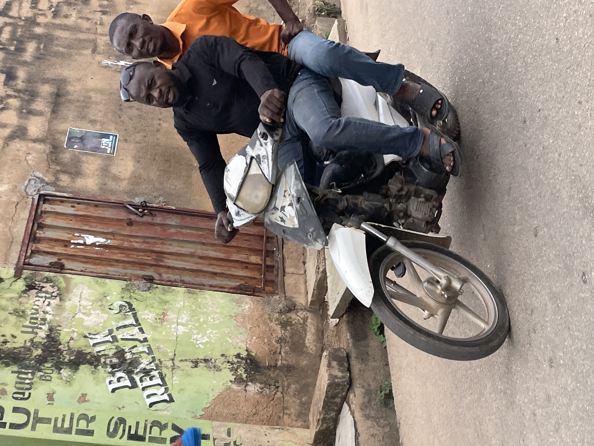 Two dark-skinned men are riding a motorcycle in Jos, Nigeria. The driver is wearing a black long-sleeved shirt, blue jeans, sunglasses on his head, and black sandals. The passenger, located behind him, is wearing an orange shirt and blue jeans with black sandals. They are on a paved road next to a deteriorating building with a green advertisement written on it, as well as a partially rusted metal door and a flyer on the wall. The motorcycle appears to be in a state of disrepair, with a partially damaged headlight.