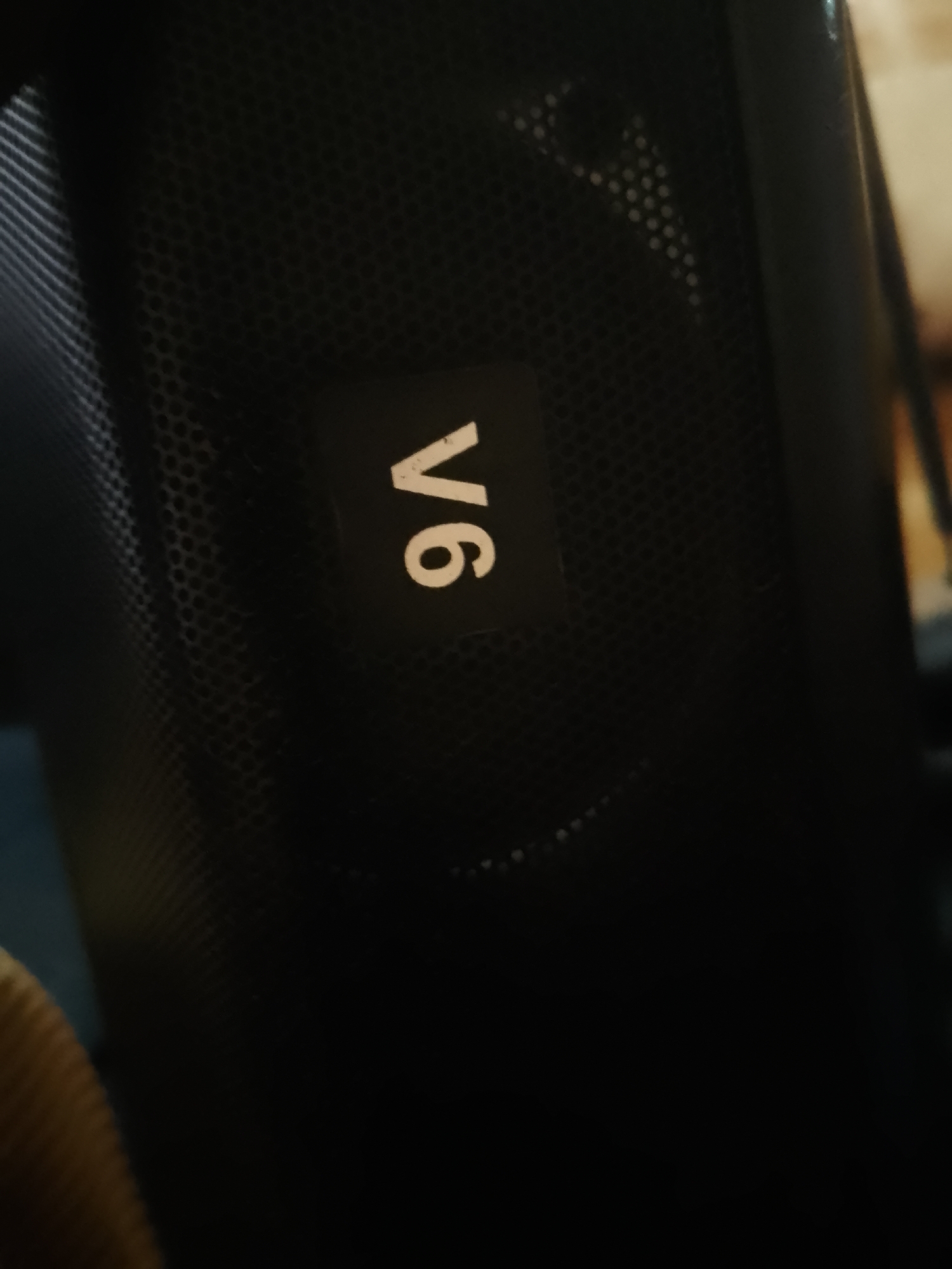 The image shows a close-up of a black object, possibly an electronic device, with a textured surface. A white emblem with the text "V6" is prominently displayed on the object. The lighting is dim, and the background is blurred, suggesting the object is indoors. The location in Serti, Nigeria, is not visually represented in the image.