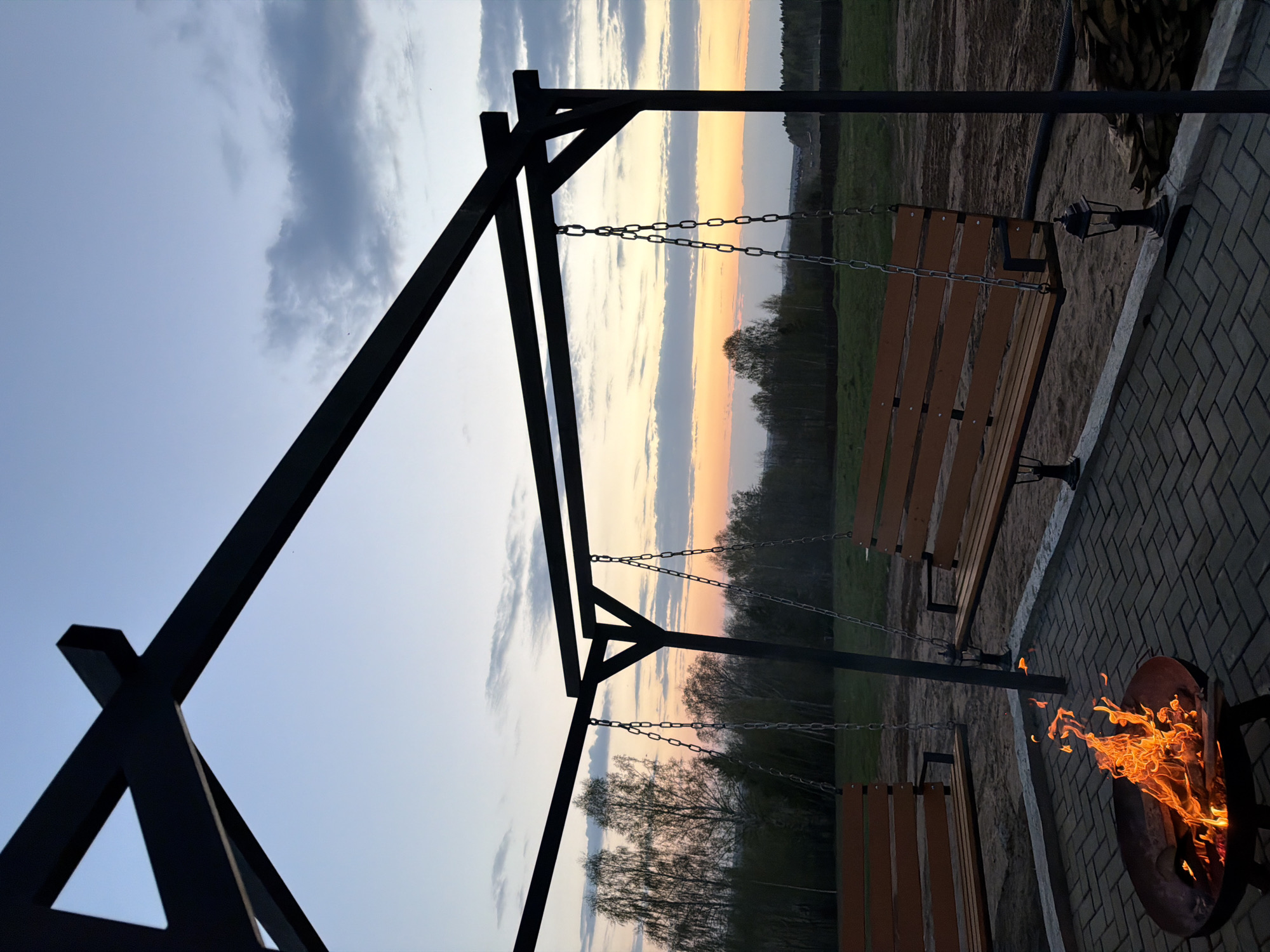 The image depicts an outdoor scene at dusk, possibly in Kamennoye, Russia. In the foreground, a black metal swing set with two wooden benches hangs suspended by chains. Adjacent to the swing set, there is a fire pit with flames clearly visible. The ground is paved with brick. In the background, a line of trees is silhouetted against a sky with horizontal bands of color, indicating a sunset or sunrise. The overall environment suggests a peaceful, possibly recreational, setting. There are no people visible in the frame.
