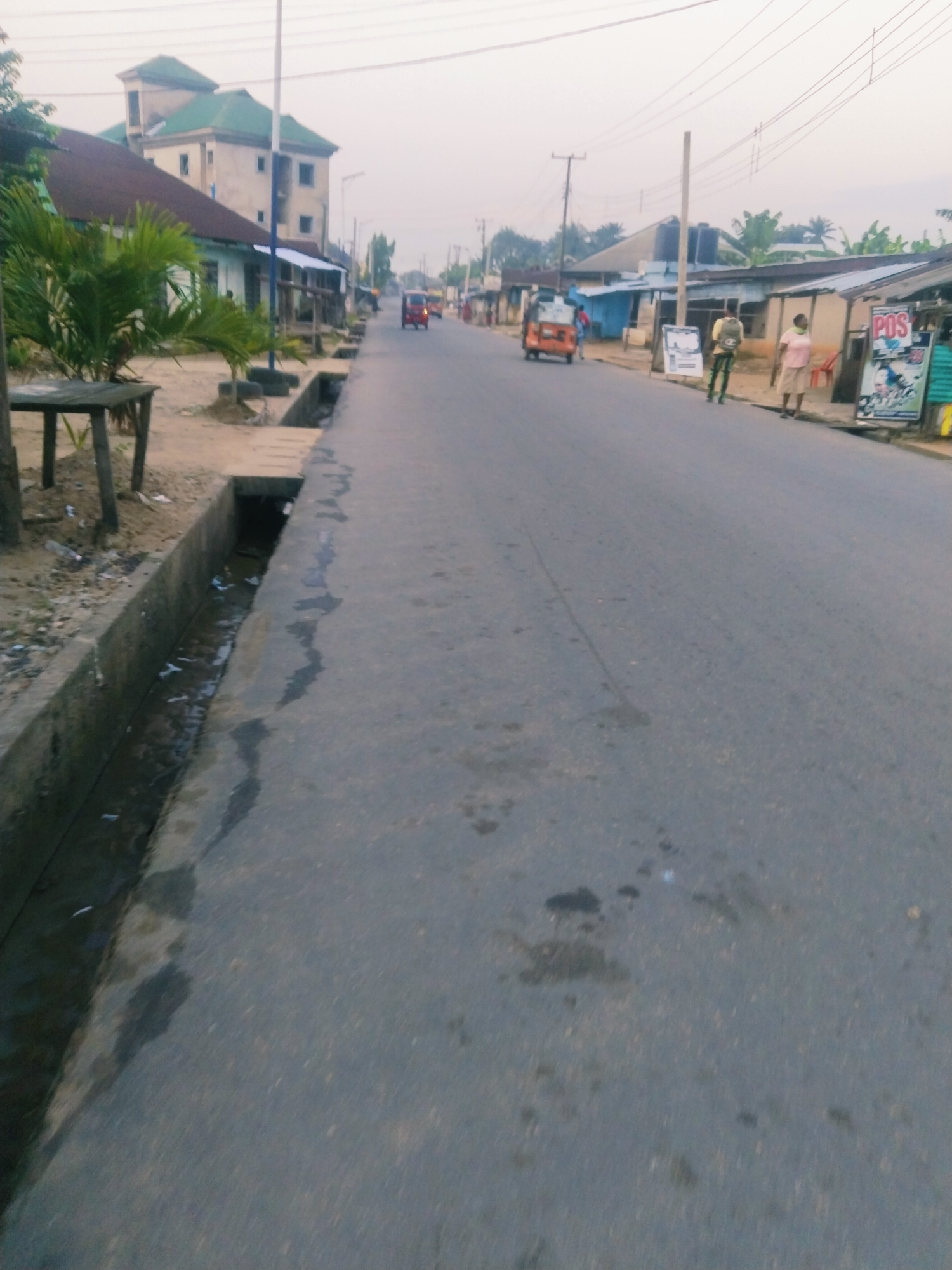 The image depicts a street scene in Uyo, Nigeria, likely during the early morning or late afternoon due to the subdued lighting. The street is paved with asphalt and appears to be a two-lane road with a concrete drainage channel running along the left side. Several buildings are visible along the right side of the road, including what appears to be a commercial establishment with a "POS" sign. Two individuals, a man and a woman, are standing near this establishment. Further down the road, a tricycle, commonly known as an "okada" or "keke," is visible, along with other indistinct figures and vehicles in the distance. A tall utility pole with multiple wires runs parallel to the road. The environment suggests a mix of residential and commercial activity in a developing urban area.