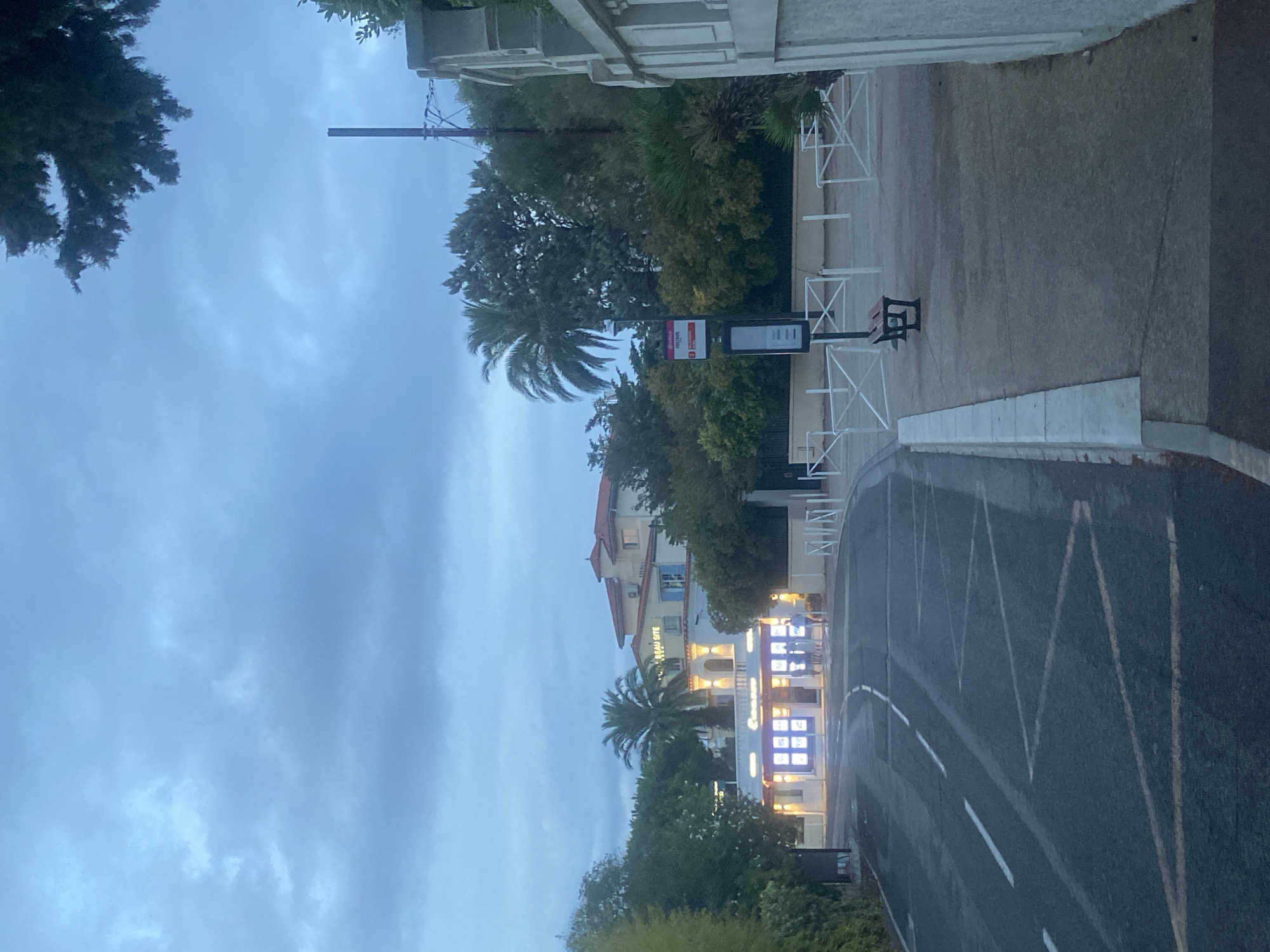 The image depicts a street scene in Antibes, France, likely near the coast given the presence of palm trees and the Mediterranean architectural style. The photograph is taken at dusk, with a cloudy sky casting a subdued light over the environment.
In the foreground, a two-lane asphalt road is visible with white lane markings and a zigzagging yellow line, suggesting a potential no-passing zone or a traffic calming measure. A paved sidewalk runs along the right side of the road, bordered by a low concrete curb. On the sidewalk, a bus stop is present, indicated by a red and white sign with a route number and the word "ROUTE." A black metal bench with a wooden seat is positioned near the bus stop. A white, decorative metal fence separates the sidewalk from a property to the right, which has a textured white wall.
Further down the road, a brightly lit building with illuminated storefronts is visible. The building features large display windows showcasing what appear to be real estate listings, as indicated by text and images within the windows. Above the storefronts, the building has an upper level with a salmon-colored tiled roof and light blue shutters. A prominent palm tree stands in front of this building. Text on the upper facade reads "BEAU SITE." Three individuals, two appearing to be adults and one possibly a child, are standing in front of the building's entrance.
The background consists of lush green trees, including more palm trees, and the cloudy, pale blue sky. The overall atmosphere is serene, with the illuminated building providing a focal point against the darkening twilight. The scene suggests a quiet evening in a residential or commercial area of Antibes.