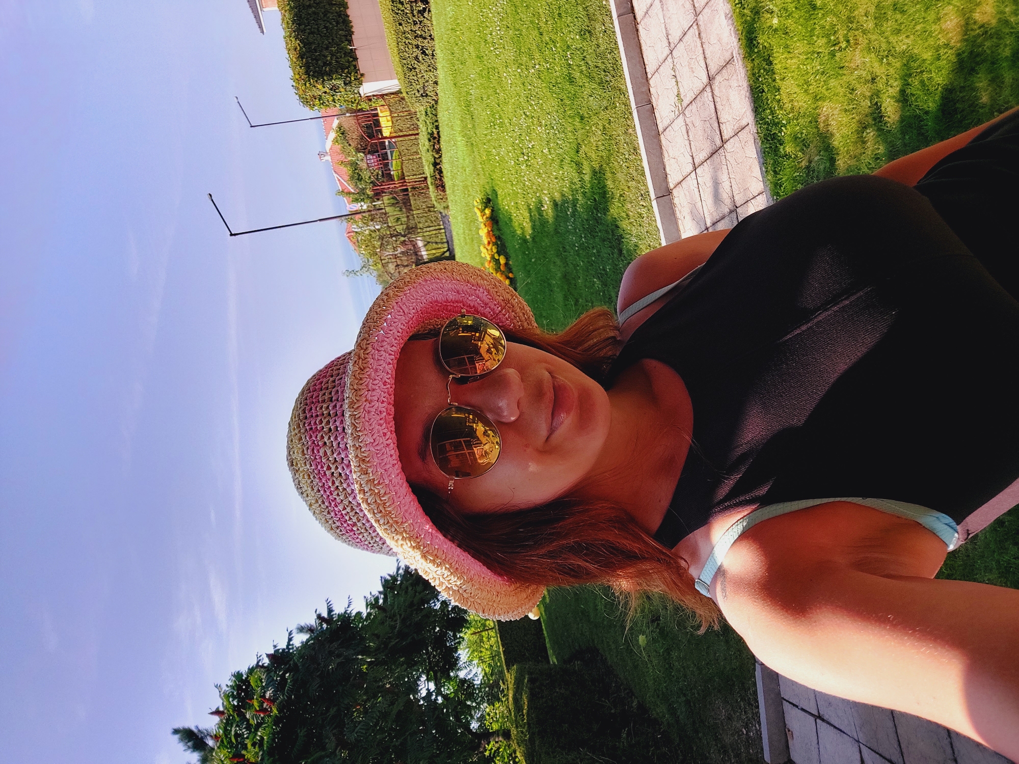 A young woman wearing a pink and yellow straw hat and sunglasses takes a selfie outdoors. She has long brown hair and is wearing a black tank top. The background shows a well-maintained lawn with a paved walkway and hedges. In the distance, there are houses with orange tile roofs, streetlights, and trees. The sky is clear and blue. The scene appears to be in a residential area or a park.