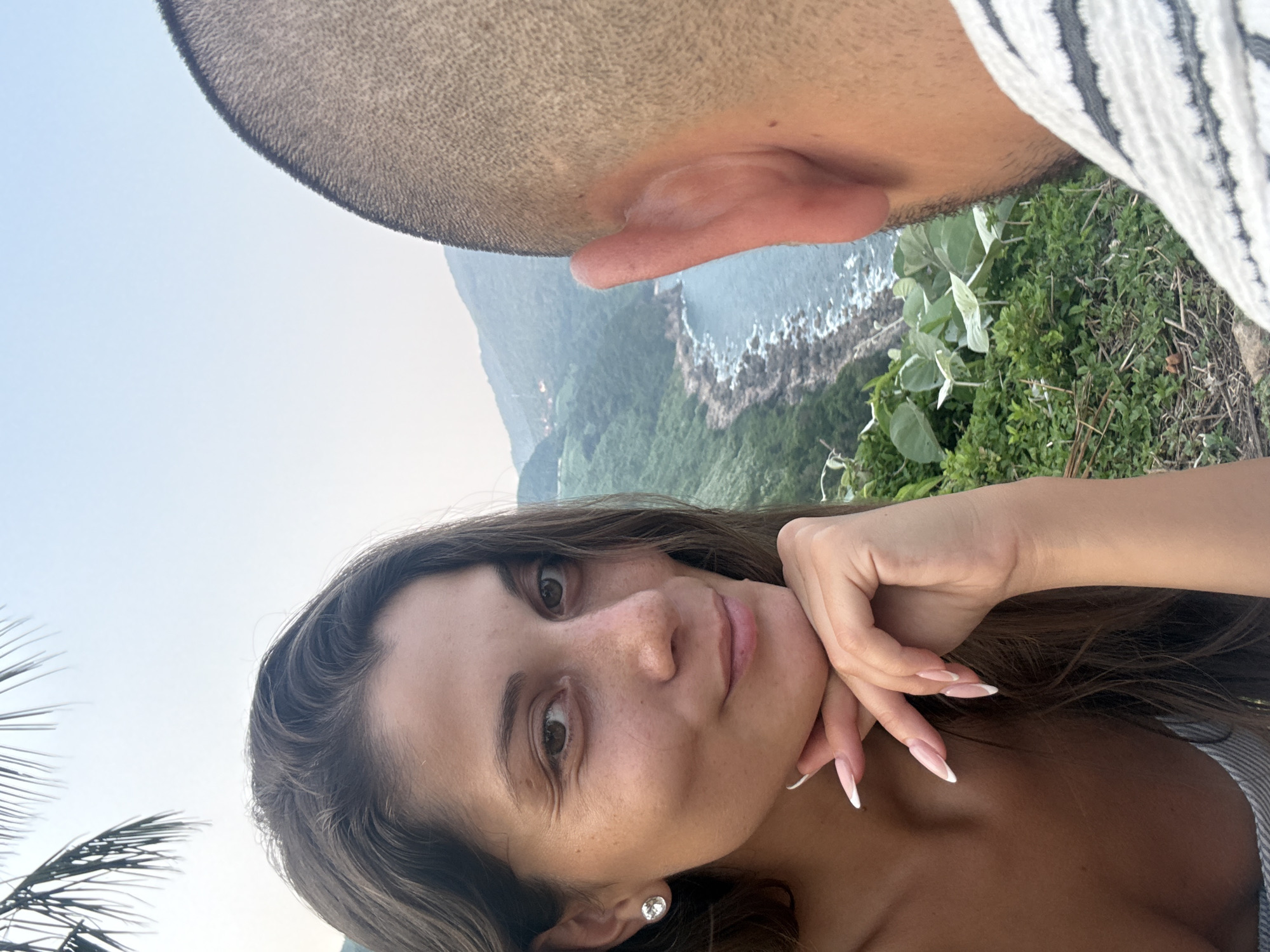 A woman is posed for a selfie, cradling her face with her hand. She has long brown hair, brown eyes, and fair skin. She wears a small earring. In the background, another individual with a shaved head and part of their striped shirt is visible. Behind them, a coastal landscape with rocky cliffs, lush greenery, and the ocean can be seen. Based on the scenery, the location appears to be in the area of Đà Nẵng, Vietnam, characterized by coastal cliffs and vegetation.