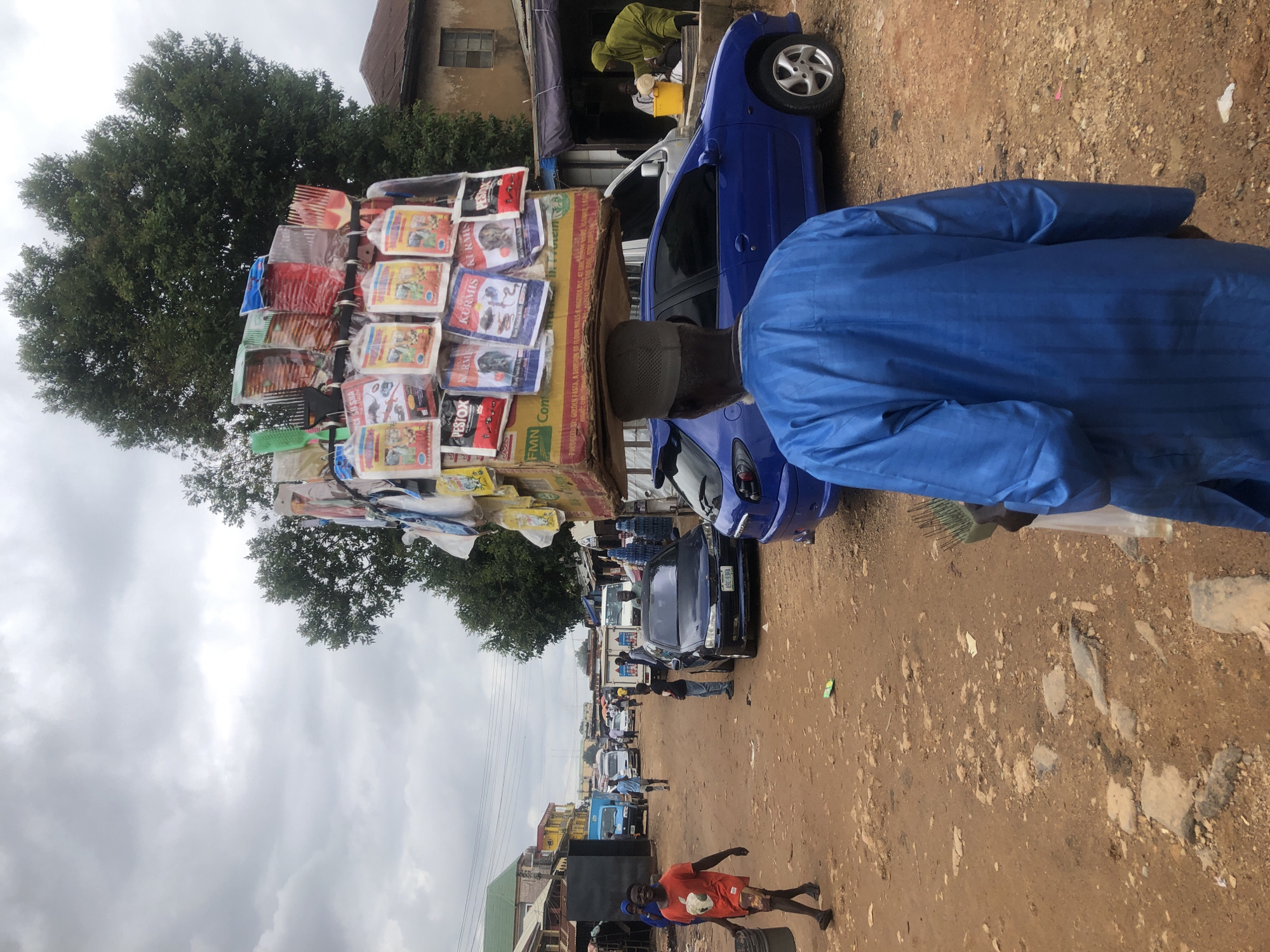 In the city of Jos, Nigeria, a man carries a large load of bagged goods balanced on his head. The goods appear to be food products, possibly pet food, as indicated by the imagery on the packaging, with brands like "Pestox" and "Kurmis" visible. He is walking on a dusty, dirt road lined with market stalls and buildings. Other people are present, including a man in an orange shirt carrying a bucket and a woman in a green hooded garment attending to a yellow bucket. Several vehicles are also in the scene, including a blue Peugeot car and other vans and cars in the background, suggesting a busy marketplace. The sky is overcast, hinting at a cloudy or possibly rainy day. The scene depicts a typical bustling market environment in Jos, showcasing local commerce and daily life.