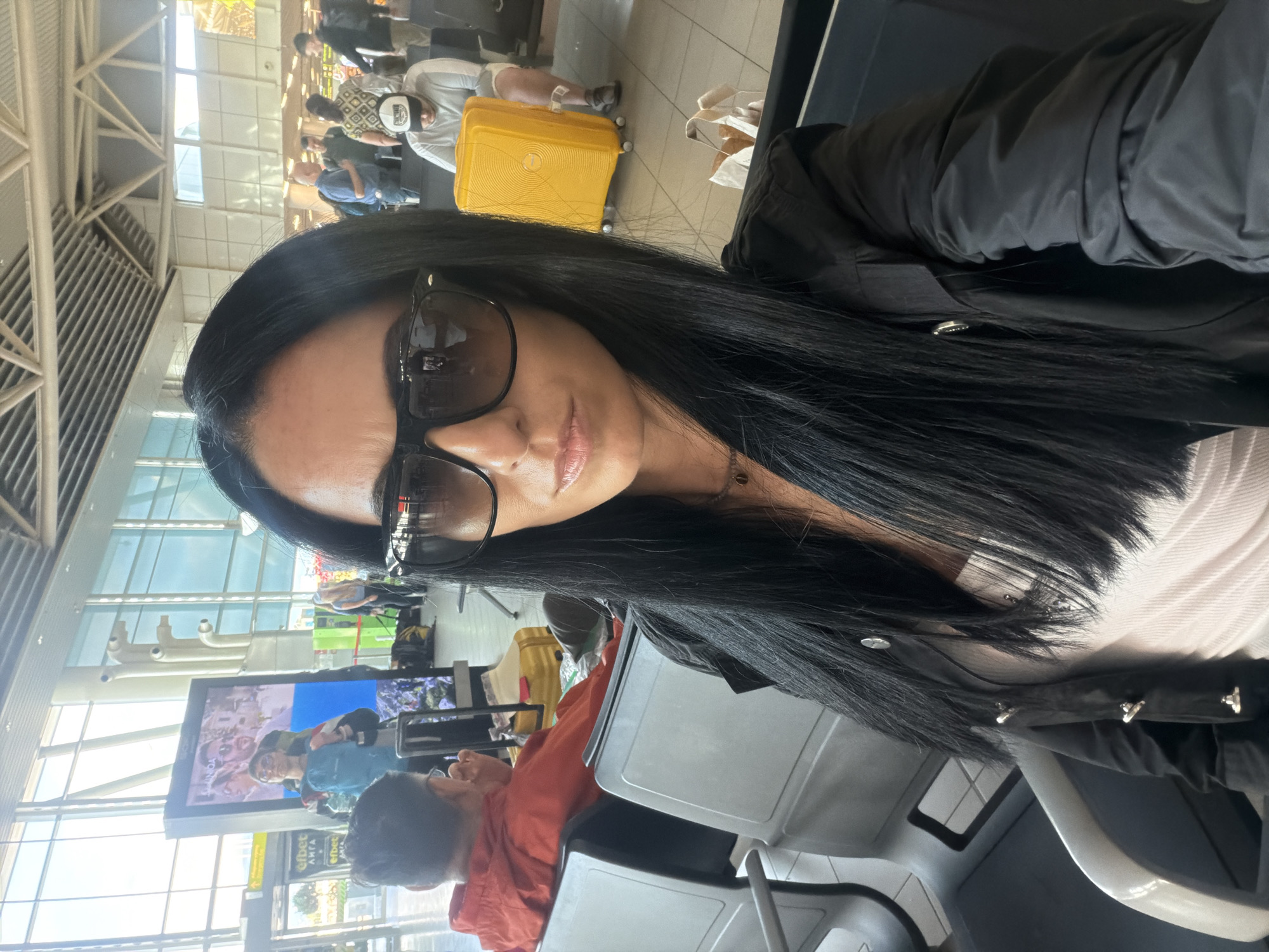 A woman with long, dark hair and sunglasses is taking a selfie in an airport waiting area. She is wearing a black jacket over a white top. In the background, other people are sitting and standing, and luggage is visible, including a bright yellow suitcase. The environment appears to be inside a modern airport terminal with large windows and a high ceiling. There is a large digital display screen in the background showing an advertisement. The scene is likely Sofia Airport, Bulgaria.