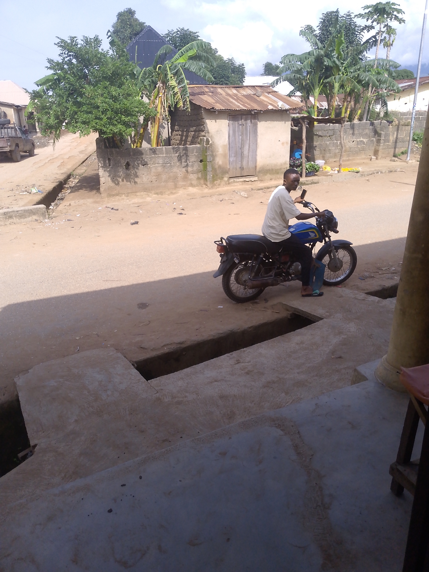 In Serti, Nigeria, a man in a light-colored t-shirt and dark pants sits astride a blue and black motorcycle. The motorcycle is parked on a dirt road in front of a concrete structure with a ditch running along its edge. Behind the man and motorcycle, there are several buildings with corrugated metal roofs and one with a wooden door. Lush green trees, including banana trees, are visible in the background. The environment appears to be a rural or semi-urban area, with the ground covered in dirt and scattered debris.