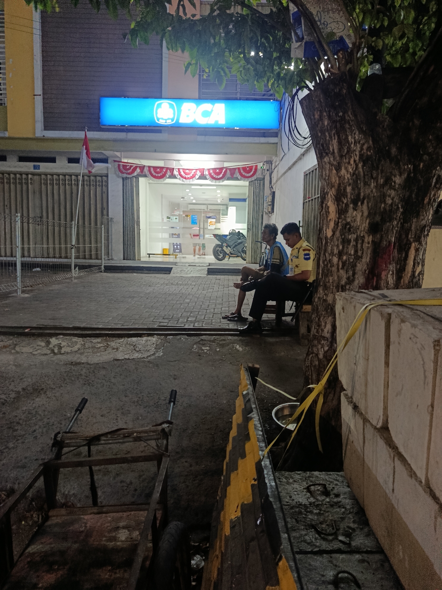The image depicts a street scene at night in front of a Bank Central Asia (BCA) branch in Surabaya, Indonesia. A bright blue sign with the BCA logo and lettering illuminates the building's facade, which also features red and white festive bunting. To the left of the entrance, a closed metal security gate and an Indonesian flag on a pole are visible.
In the foreground, a rusty metal handcart with two wheels sits on a paved area. A large tree with rough bark is prominently positioned on the right side of the image. Two men are seated on a wooden bench next to the tree. One man, appearing older, is wearing a blue shirt and shorts. The other man, a security guard, is in uniform, a yellow shirt with dark pants, and appears to be holding a mobile phone. Inside the bank, a motorcycle is parked, and another person is partially visible near the entrance. The ground is paved with cobblestones and concrete, with some areas appearing wet. A large concrete structure with yellow caution tape wrapped around it is on the far right, partially obscuring the view of the building. The overall lighting suggests it is nighttime, with artificial light sources from the bank and streetlights illuminating the scene.