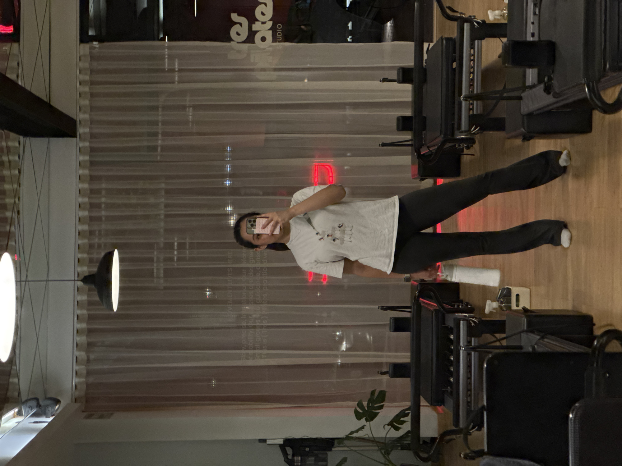 A young woman is captured in a mirror selfie within what appears to be a Pilates studio in Puebla, Mexico. She stands casually, holding a smartphone that obscures part of her face, and is dressed in a white t-shirt, dark leggings, and white socks.
The studio features a light wooden floor and several dark-colored Pilates reformer machines. In the background, sheer, light-toned curtains cover a wall, through which reflections and a red neon sign are visible, suggesting it is evening or night outside. A potted plant with large leaves is in the lower foreground, and overhead, modern light fixtures illuminate the space. Other items visible include a white water bottle and a wall-mounted dispenser.
Visible text in the image includes "MOVEMENT LIVES HERE," "STUDIO," and partially readable words like "pilates" and descriptions about "Slow, precise, intentional Our eform," and "Small groups." The overall atmosphere is that of a modern fitness facility, likely at the end of a workout session.