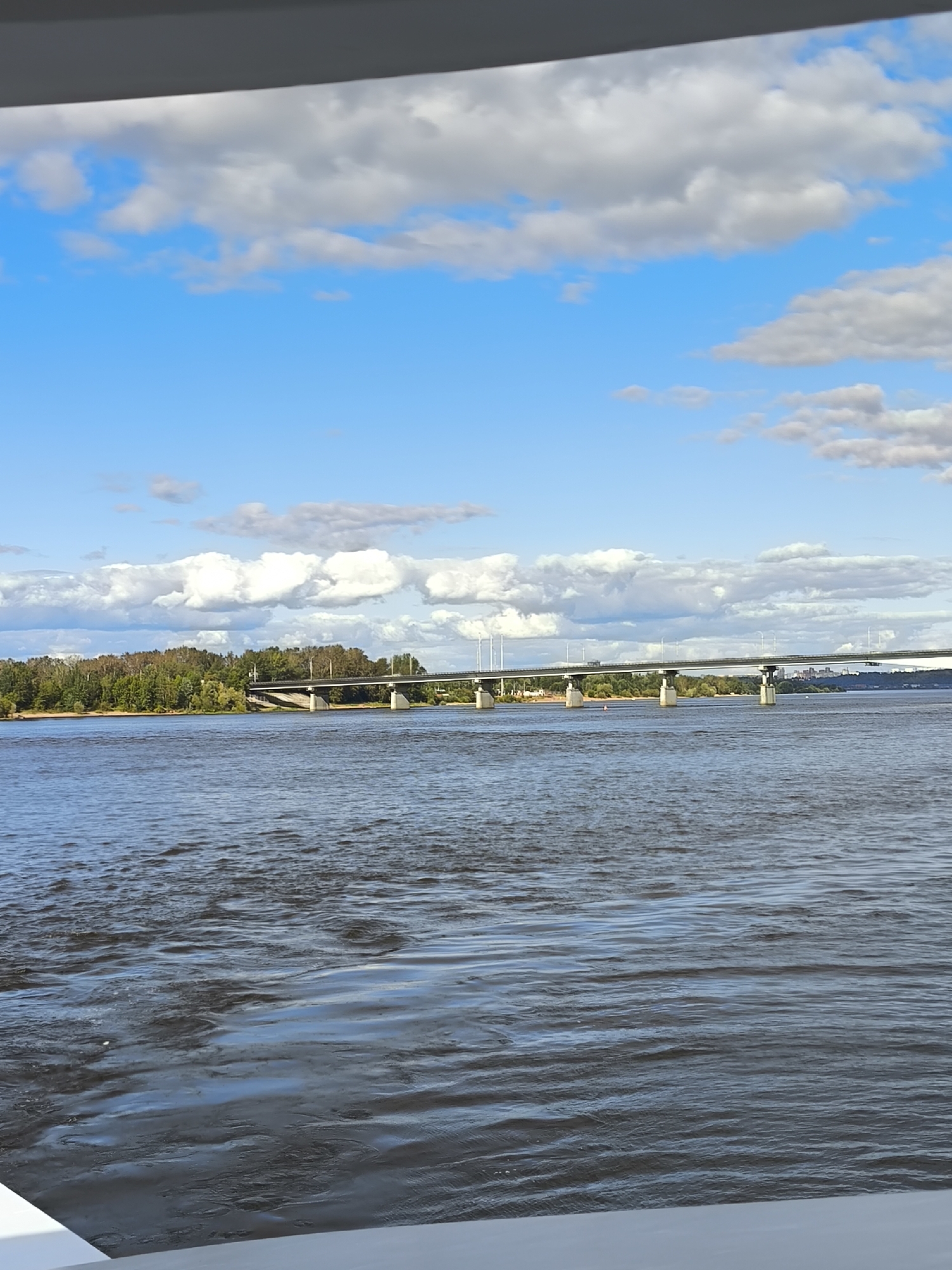 An exterior, horizontal, eye-level shot depicts a wide, choppy river with a long, multi-span concrete bridge crossing it. The bridge is supported by numerous concrete piers and has a road surface lined with streetlights. On the far bank, there is a dense, green forest. The sky is bright blue with scattered white and gray clouds. In the foreground, a white railing is partially visible. The scene is likely the Kama River in Perm, Russia, given the presence of a large bridge spanning a wide body of water with a forested bank, characteristic of the city's landscape.