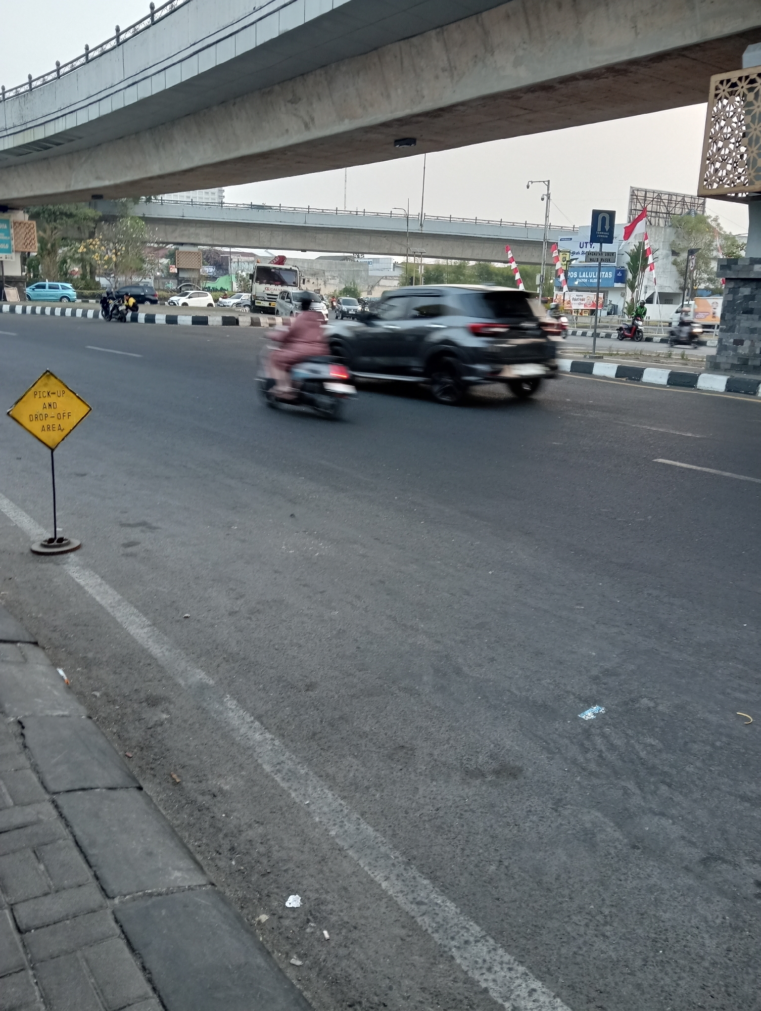 In Sinduadi, Indonesia, an image captures a view of a street with an overpass. A yellow, diamond-shaped sign is prominently displayed on a metal pole at the left side of the road, reading "PICK-UP AND DROP-OFF AREA". The road surface is asphalt, with white lane markings visible. Cars and a motorcycle are in motion on the road, with motion blur indicating they are moving. The environment suggests an urban setting with a bridge overhead and buildings in the background. The sky is overcast.