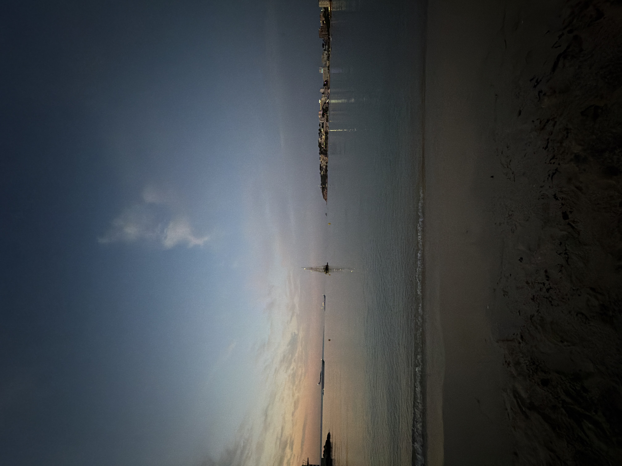 The image depicts a serene twilight scene at a beach in Calvià, Spain. The foreground is a dark, sandy beach with gentle waves lapping the shore. In the middle ground, the calm sea reflects the fading light of the sky, which transitions from warm orange and pink hues near the horizon to a deep blue overhead. Several boats are visible on the water, including a sailboat with its mast prominently featured and a larger vessel further in the distance. On the far right of the image, a distant coastline is illuminated by the lights of buildings, suggesting a town or city settlement. The overall atmosphere is tranquil and peaceful, characteristic of an evening by the sea.