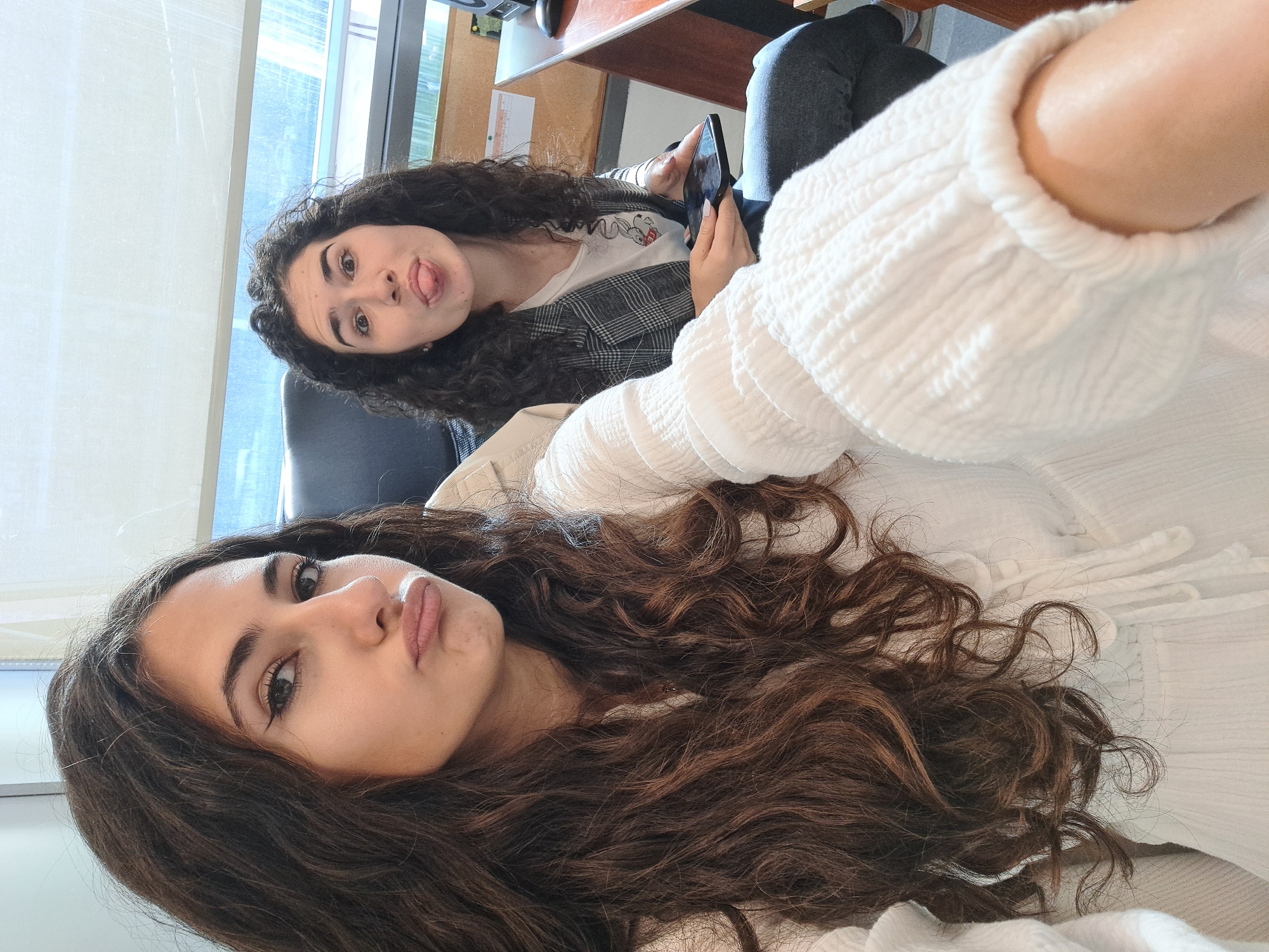 Two young women are in an indoor setting. The woman closest to the camera has long, dark, curly hair and is wearing a white top. Her lips are pursed. The other woman has dark, curly hair and is sticking out her tongue. She is wearing a plaid jacket over a white t-shirt and holding a black smartphone. They appear to be taking a selfie. The background includes a window with blinds, a desk, and a chair. The location is most likely Sofia, Bulgaria.