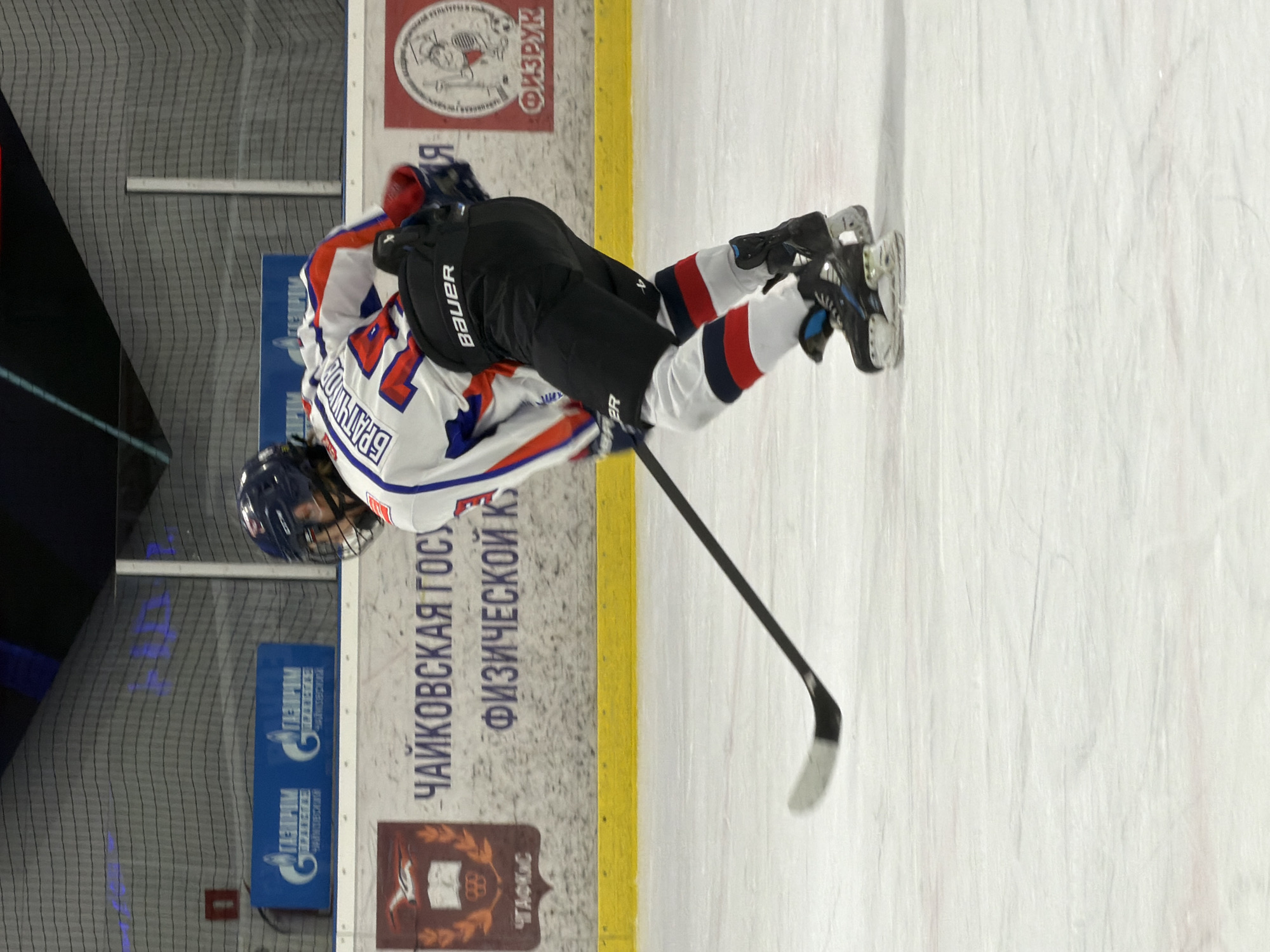 A hockey player wearing a blue helmet, a white jersey with blue and red accents, black pants with a "Bauer" logo, and white and blue skates is on the ice in a rink. The player holds a black hockey stick. The boards of the rink are visible in the background, displaying "ЧАЙКОВСКАЯ ГОС" and "ФИЗИЧЕСКОЙ К". Logos and other markings are also on the boards. The image suggests the location is Chaikovsky, Russia.
