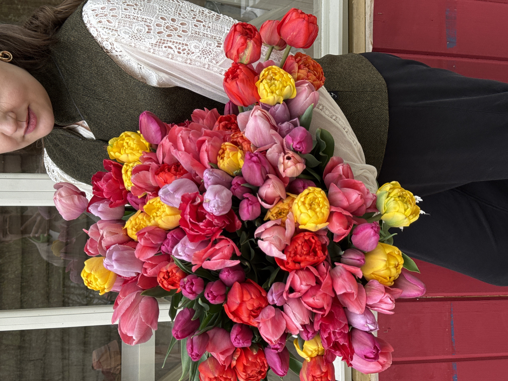 A woman in Sapronovo, Russia, is holding a large bouquet of tulips in various colors, including red, pink, yellow, and purple. She is wearing a white lace top and a dark green vest. Her dark pants are also visible. The background shows a white window frame and a red painted wall.
