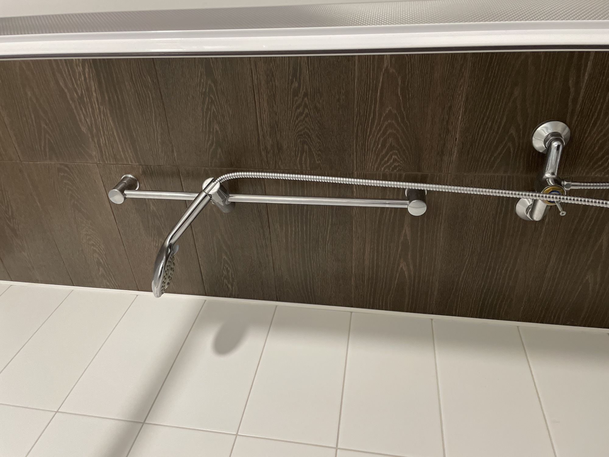 An indoor shot shows a showerhead and faucet against a backdrop of dark wood-grained tiles and white rectangular tiles. The showerhead is attached to a silver adjustable bar. The faucet has a silver handle with a central knob. The scene is located in Sinyavino, Russia, within a bathroom. There are no people present in the image.