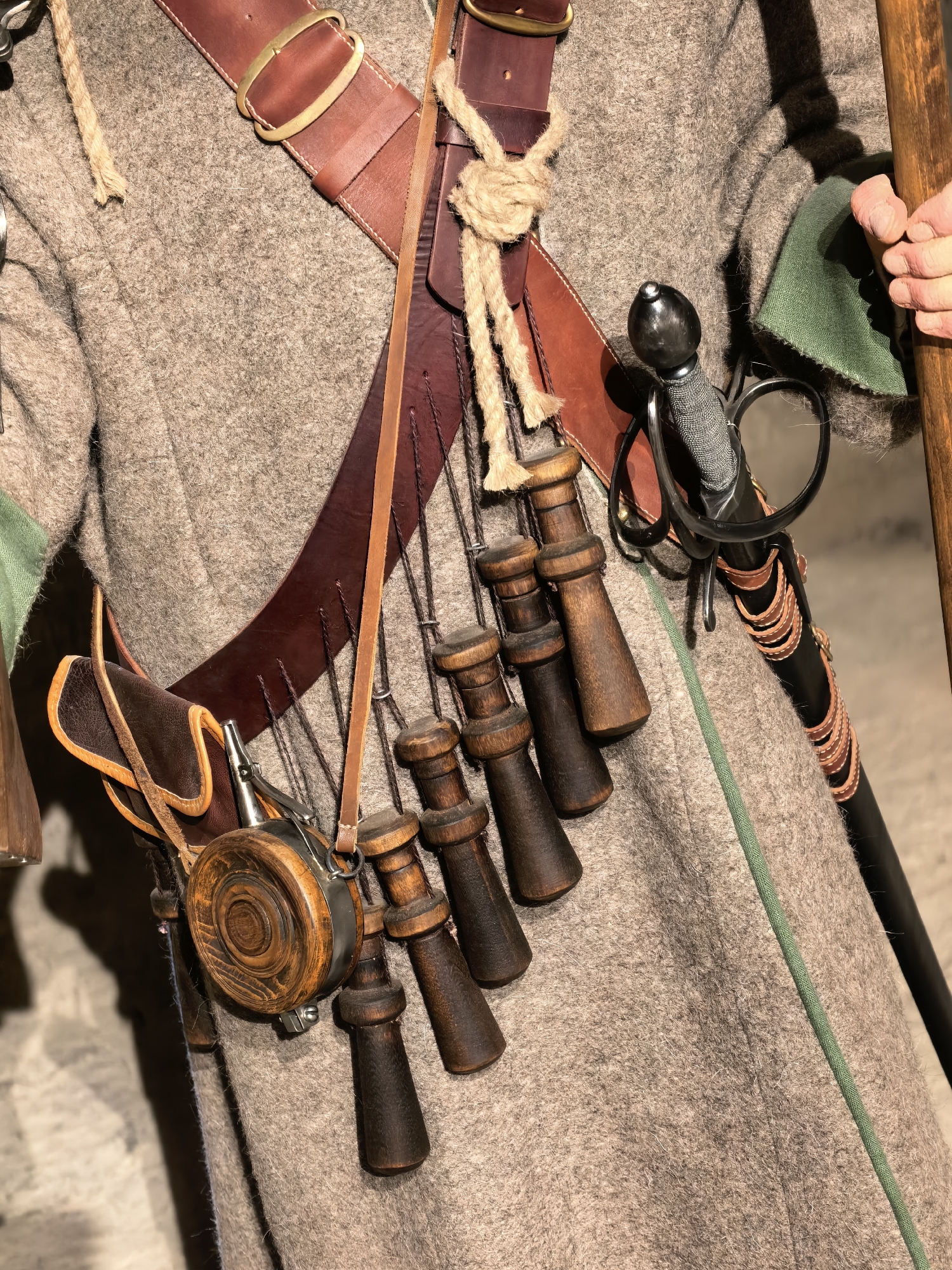 The image shows a close-up, waist-up view of a person dressed in historical clothing, likely a reenactor, in Staraya Ladoga, Russia. The individual is wearing a rough, tan woolen coat with green fabric visible at the cuffs. A wide leather belt, secured by a brass buckle, is worn over the coat. Suspended from the belt are several items: a leather pouch, a small round flask made of wood and metal, and a series of wooden powder horns. A sword with a dark hilt and metal guard is sheathed in a black scabbard adorned with leather straps. A wooden staff or spear is visible on the right side of the frame, held by a hand. The background is a blurred, indoor setting.