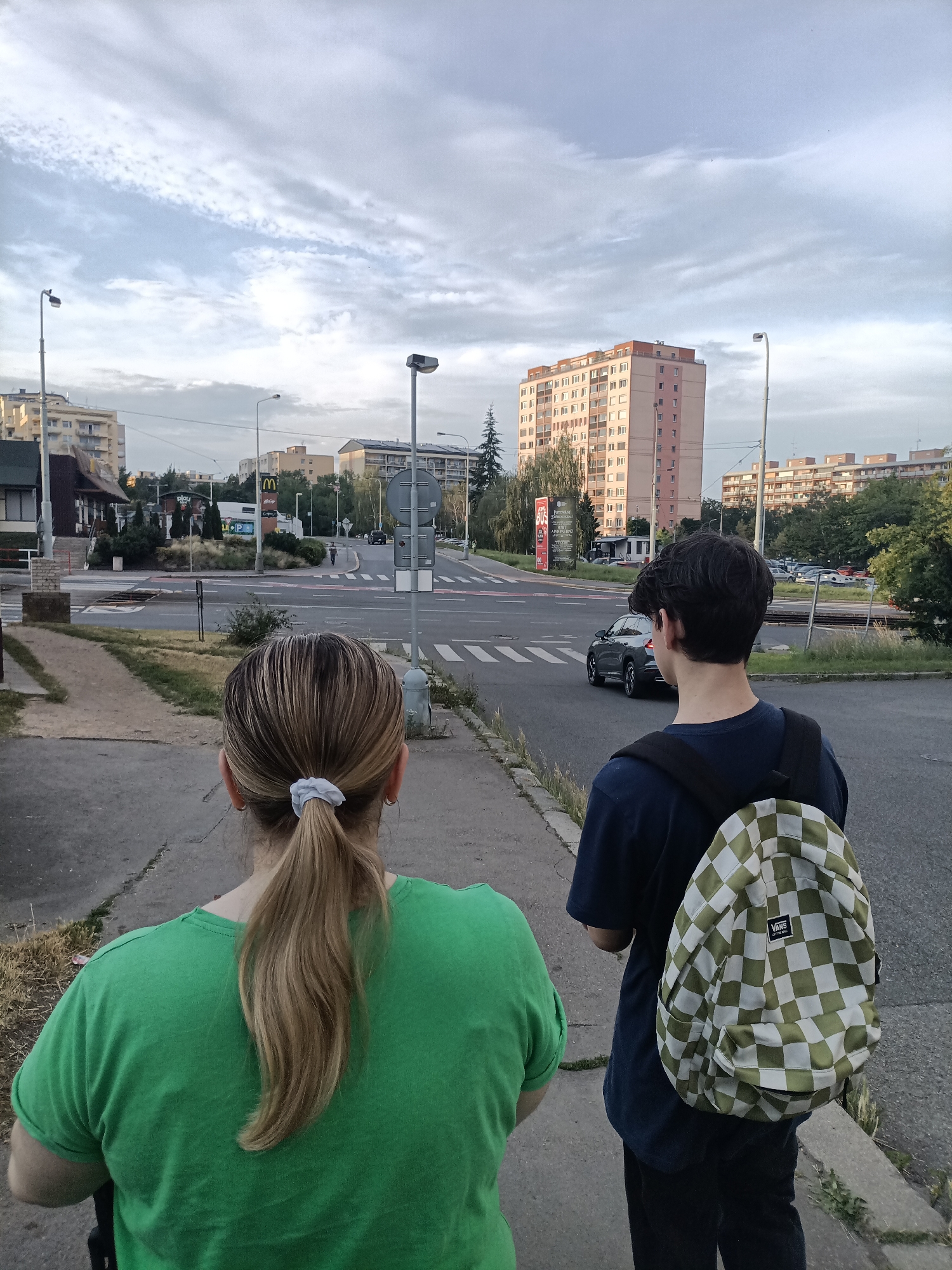 The image shows two people, a woman and a young man, standing on a sidewalk facing a street intersection in a city. The woman, seen from behind, has blonde hair pulled back in a ponytail and is wearing a green t-shirt. The young man, also seen from behind, has dark hair and is wearing a dark blue t-shirt and a checkered backpack. They are looking towards the intersection where a dark gray car is driving. In the background, there are several multi-story apartment buildings, streetlights, and businesses, including a McDonald's restaurant. The sky is partly cloudy. The scene appears to be in an urban residential area. While the exact location is not specified in the image, the presence of a McDonald's and the architectural style of the buildings suggest it could be a common urban environment in Europe, possibly within Prague, Czechia.