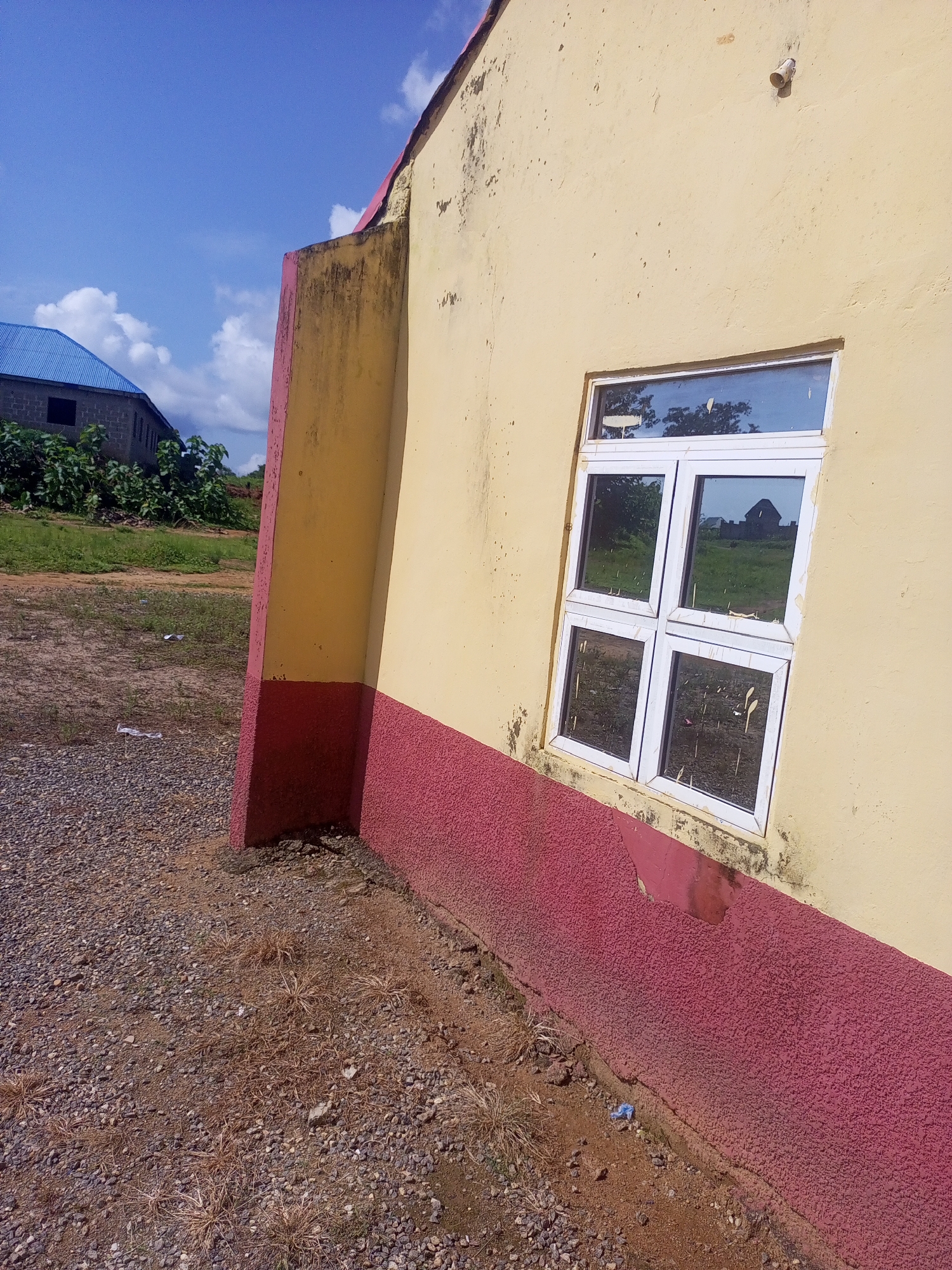 This is a medium-angle exterior shot of a building in Serti, Nigeria. The building has a yellow stucco facade with a thick red base. A white-framed window with four panes is visible on the yellow wall. The window reflects trees and a cloudy blue sky. To the left of the building, a gravel path leads to a grassy area with vegetation and another building with a blue roof. The ground is covered in gravel and dry grass. The scene appears to be a sunny day.