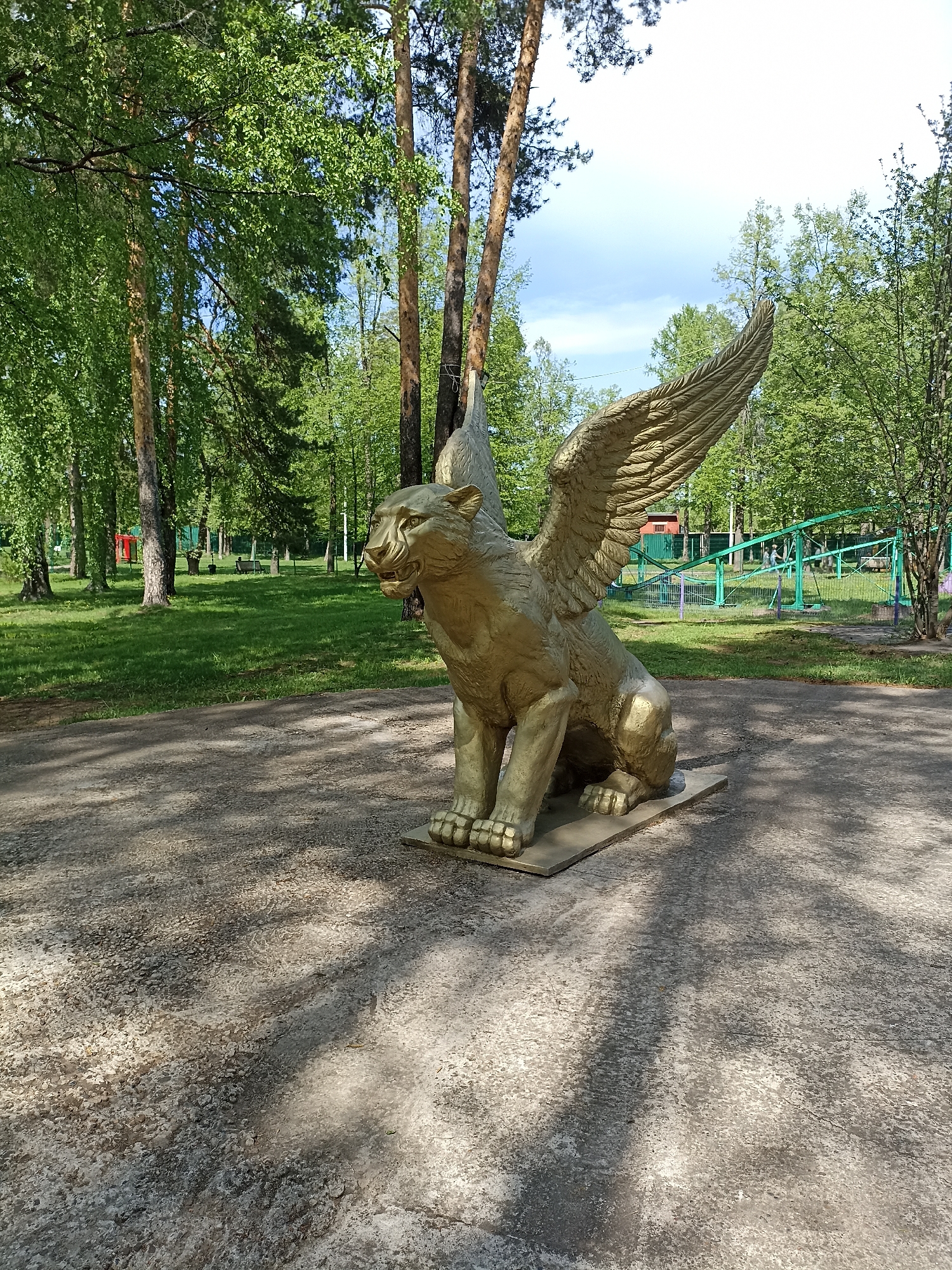 A golden griffin statue stands on a concrete path in a park in Izhevsk, Russia. The statue has the body of a lion with the head and wings of an eagle. The griffin is in a seated position, facing left. Behind the statue is a park setting with green trees, a green fence, and a playground structure. The sky is partly cloudy.