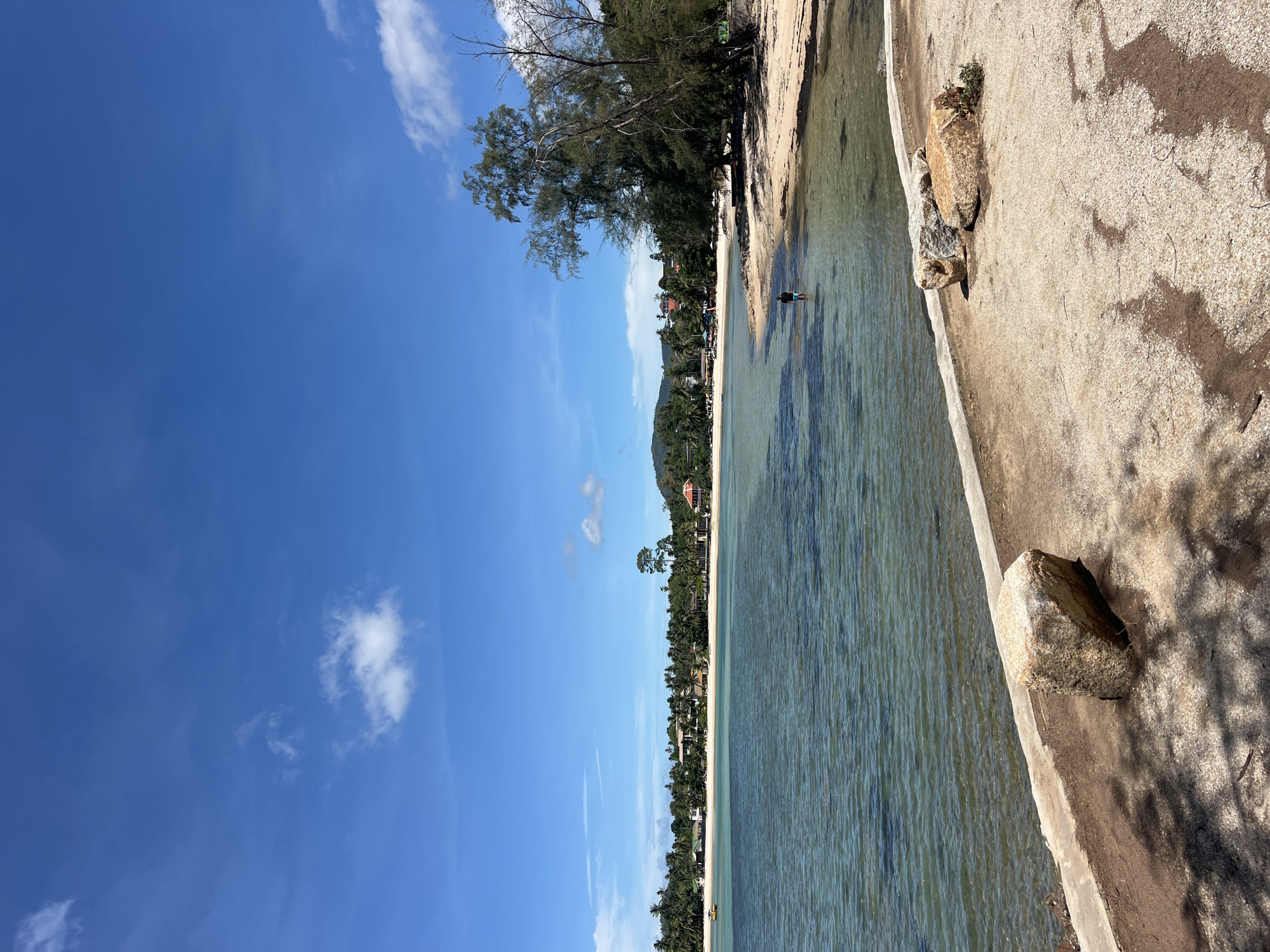 The scene is a beach in Baan Hua Thanon, Thailand, taken during the day. A concrete path runs parallel to the water's edge, marked by rocks along its edge. The water is clear and shallow, revealing the sandy bottom. A person stands in the water. Beyond the beach, trees line the shore, with buildings visible among them. A hill rises in the distance. The sky is blue with scattered clouds.