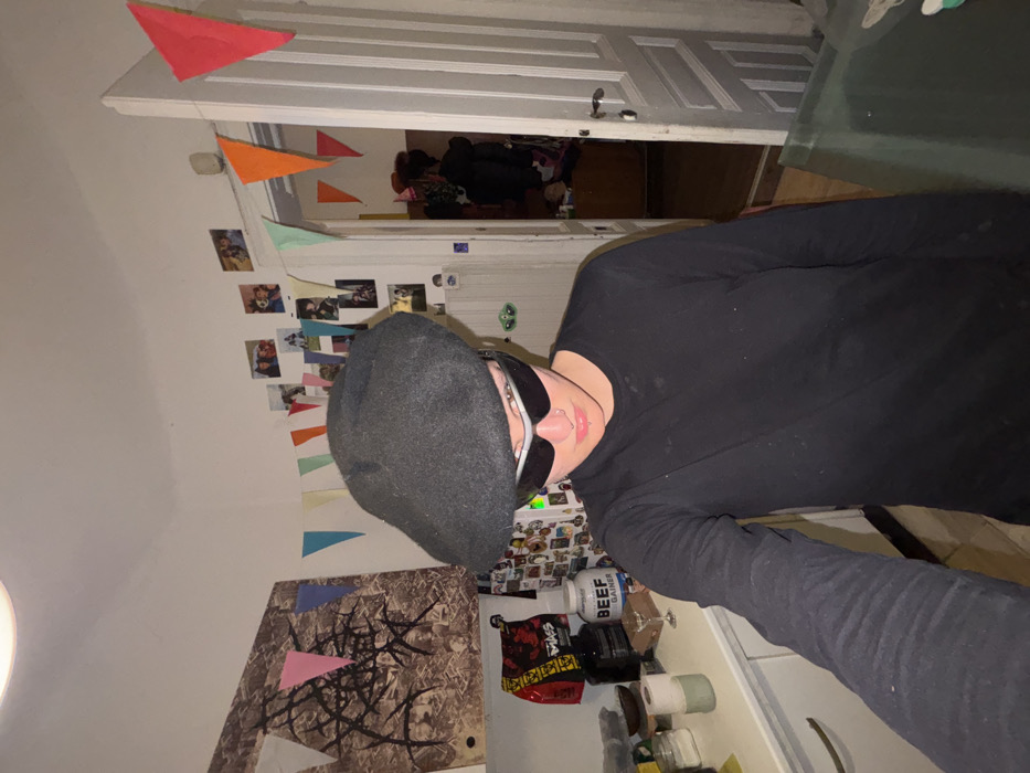 An individual is depicted indoors, taking a selfie. The setting is a domestic residence in Sofia, Bulgaria. The person, positioned centrally, wears a dark grey beret, white-rimmed dark sunglasses, and a long-sleeved black shirt. They have a septum piercing and a philtrum piercing. Their right arm is extended, holding the device used to capture the image.
The background on the left features a white wall with a large abstract artwork showing dark, branching forms on a textured brown surface. Below this, a white kitchen counter holds various items including a red and black bag (possibly marked "MASS" and "CREATINE"), a white tub labeled "BEEF GAINER," a black appliance, several small containers, a wine glass, and two rolls of paper. A segment of a refrigerator adorned with magnets is also visible.
The background on the right side of the room displays a white wall covered with numerous small photographs. Colorful triangular bunting, in shades of red, pink, light blue, orange, green, and yellow, is strung across the upper wall and above an open white doorway. Beyond the doorway, into an adjacent room, dark clothing items or objects are discernible, along with a brown piece of furniture and additional bunting. Overhead, an artificial light source illuminates the scene.