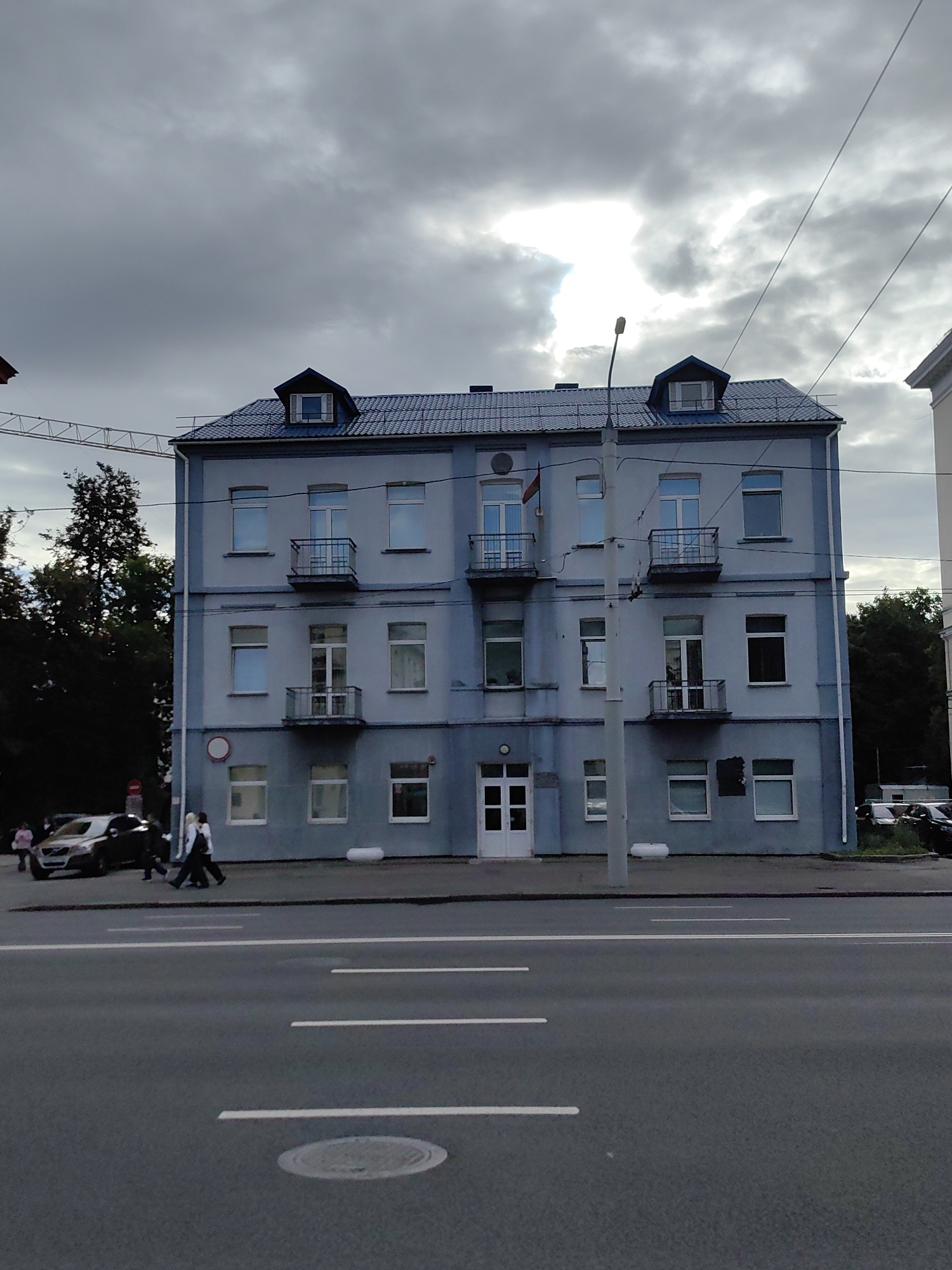 A light blue, three-story building with a dark blue, sloped roof stands on a city street. The building has numerous windows and balconies. Two dormer windows are visible on the roof. A white pole stands in front of the building. A street with lane markings and a manhole cover is in the foreground. Several cars and pedestrians are on the sidewalk near the building. Trees are visible behind the building to the left. The sky is cloudy with bright sunlight breaking through in the upper center. The scene appears to be in Minsk, Belarus, as indicated by a sign on the building that has Cyrillic lettering.