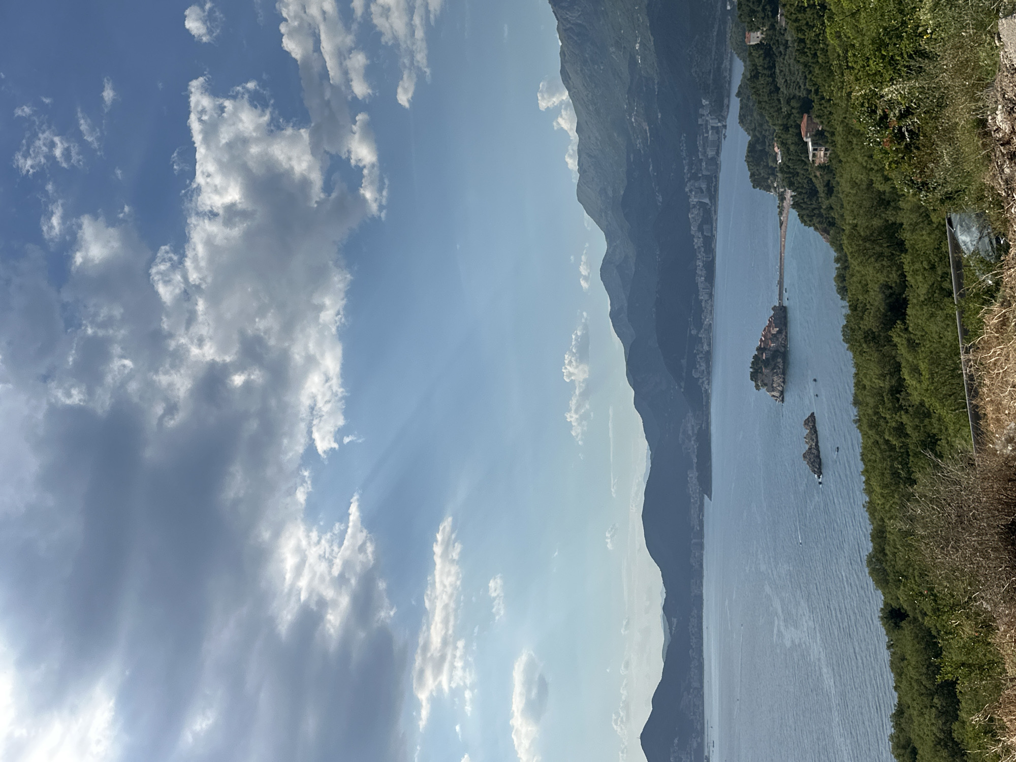 This is a vertical, eye-level, medium shot of the coastal region near Blizikuće, Montenegro, on a day with a partially cloudy sky and sunbeams breaking through. The foreground is dominated by dry, brown vegetation and some green foliage. In the mid-ground, a lush green hillside slopes down towards the Adriatic Sea. The sea itself is a deep blue, with a few small boats visible. Dominating the mid-ground is the distinctive island of Sveti Stefan, a small fortified village connected to the mainland by a narrow causeway, with its reddish-brown rooftops and surrounding green vegetation. Beyond the island, the coastline curves, revealing more green hills dotted with buildings and a distant city or town. The background consists of a range of layered, dark blue mountains, with the sky above featuring both bright blue patches and dense, gray clouds.