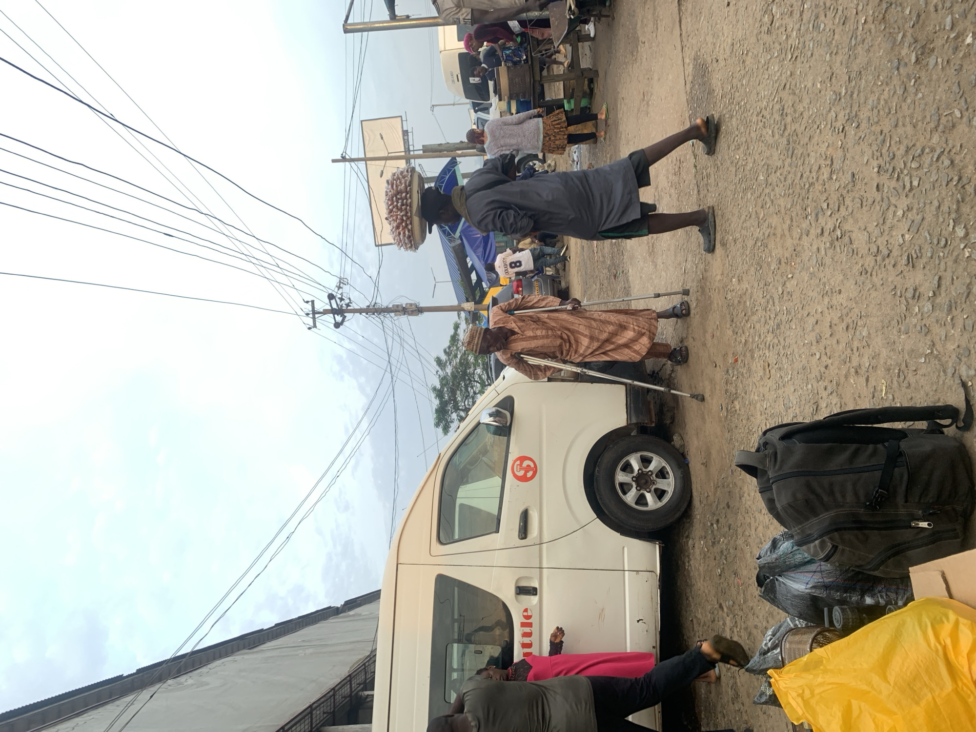 In Elelenwo, Nigeria, an outdoor scene unfolds with a white shuttle van on the left. A man in a brown, patterned robe and a cap, using two crutches for support, stands near the van. To his right, another man dressed in a dark gray robe and shorts carries a large tray of food items on his head. Behind them, several other individuals are visible, along with market stalls and various vehicles, suggesting a busy market or transportation hub. The ground is unpaved and covered with gravel and dirt, typical of the area. Power lines crisscross the sky above. In the foreground, several bags, including a yellow plastic bag and a dark backpack, are placed on the ground.
