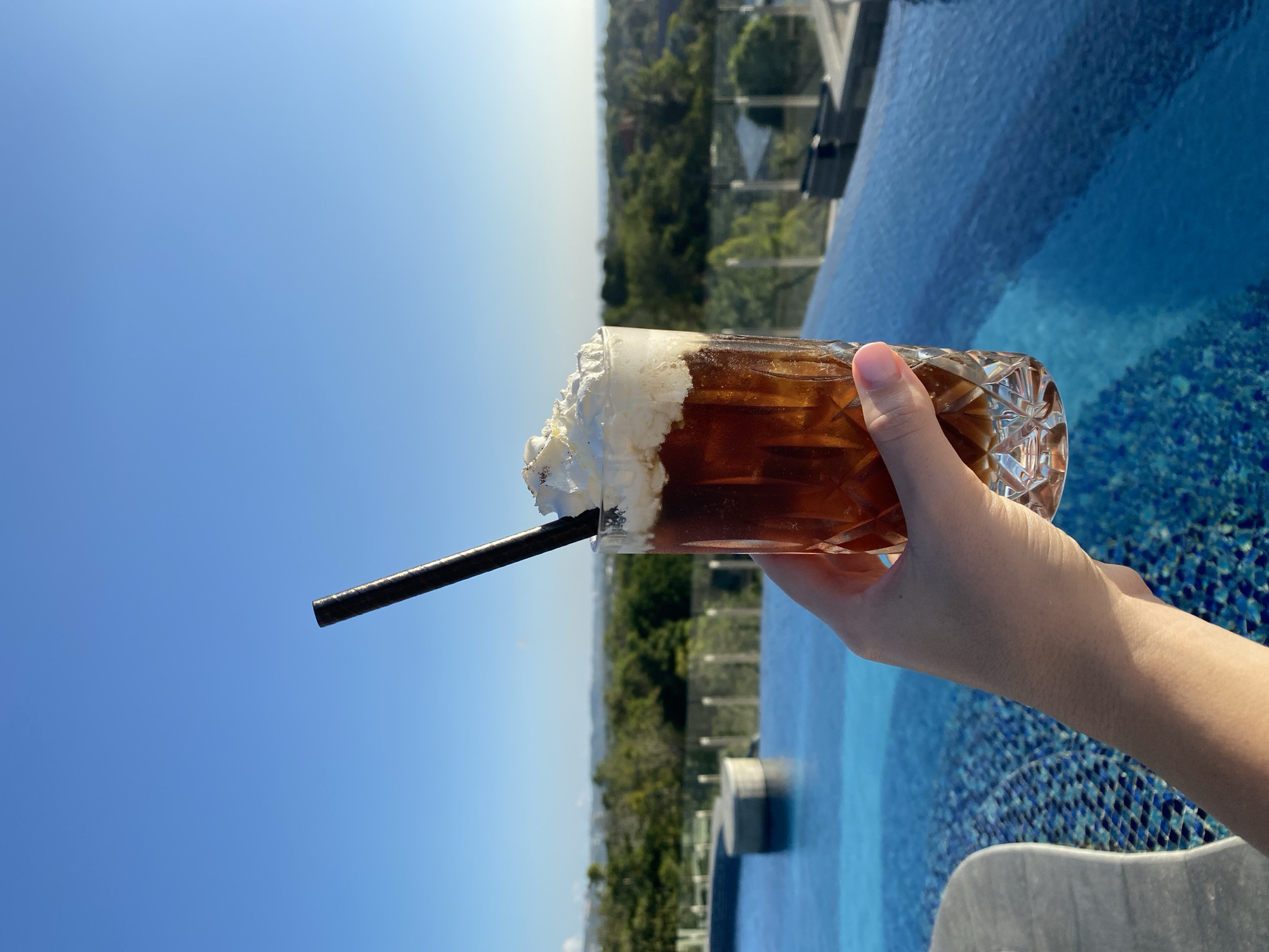 A person's hand holds a glass of iced coffee with whipped cream and a straw, positioned in front of an infinity pool and lush green landscape under a clear blue sky. The glass is clear with a diamond-cut pattern and filled with dark liquid and ice, topped with a generous amount of white whipped cream. A black straw is inserted into the whipped cream. The infinity pool's bright blue water is visible, with intricate mosaic tiles on its floor. Beyond the pool, a dense green forest stretches towards the horizon, hinting at a tropical location. The scene appears to be outdoors, possibly at a resort or a rooftop bar, overlooking a scenic vista.
