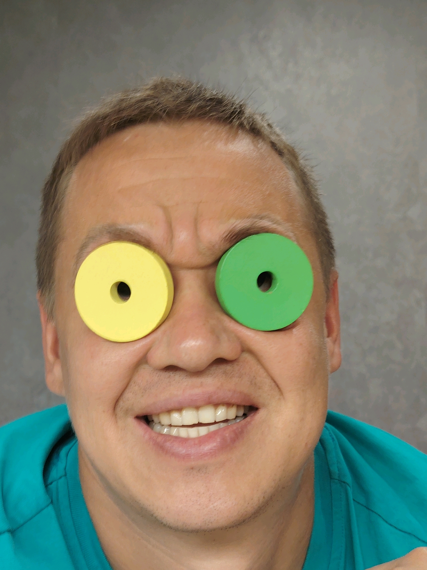 A man's face is shown from the chest up. He has short brown hair and a graying beard stubble. Two large, colored wooden discs are placed over his eyes, one yellow on the left and one green on the right. He is wearing a turquoise shirt. The background is a mottled gray. No information about the location in the city of Chaikovsky, Russia is available in the image.