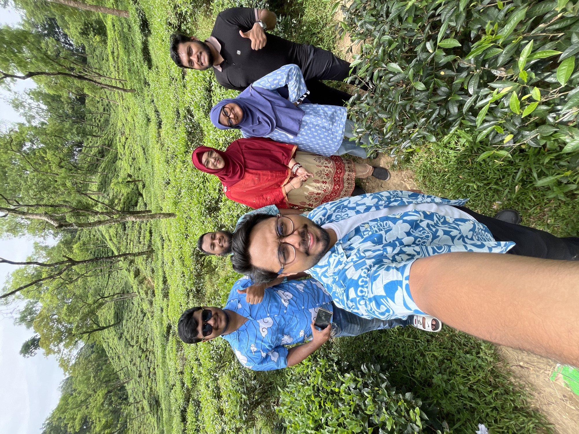 The image shows a group of six individuals posing for a selfie in a lush, green tea garden. The location is identified as Sylhet, Bangladesh, known for its extensive tea plantations.
In the foreground, the arm of the person taking the selfie is visible, wearing a blue and white patterned shirt. To their right, a man wearing glasses and a similar blue and white shirt is smiling. Behind them, from left to right, are: a man in a blue shirt with a cloud pattern, giving a thumbs-up; a man in a black polo shirt, also giving a thumbs-up; a woman wearing a red hijab and a cream and red patterned outfit; and a woman in a blue hijab and a light blue patterned top. They are all standing amongst rows of tea plants that cover rolling hills. The environment is outdoors with trees scattered on the slopes of the tea garden, and the sky appears overcast. The overall scene suggests a casual outing or visit to a tourist attraction in Sylhet.