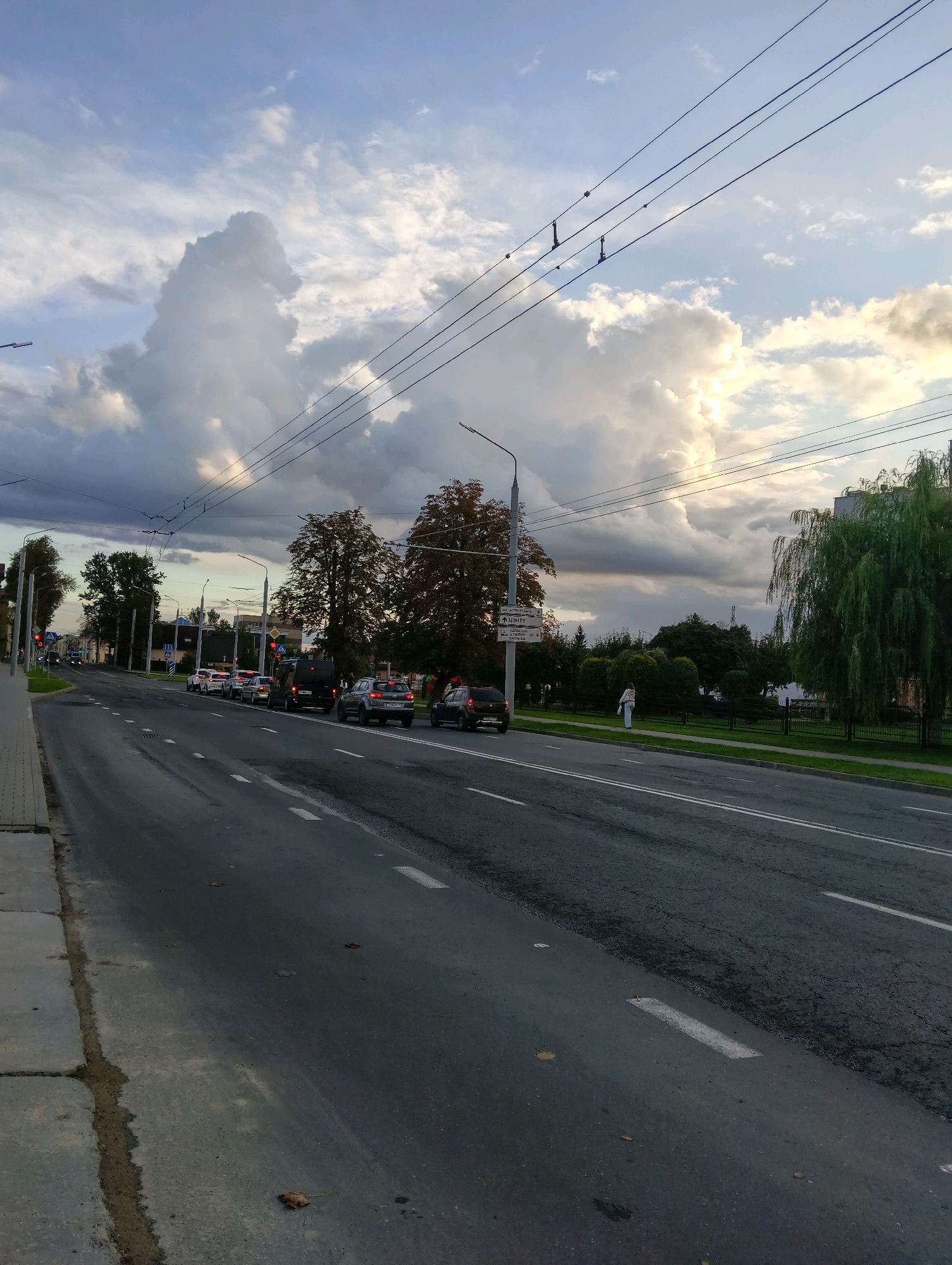 The image captures a street scene in Hrodna, Belarus, likely during late afternoon or early evening, given the sky's lighting. The perspective is from the sidewalk, looking across a multi-lane road. Several cars are visible on the road, with some in traffic. The road itself is asphalt, marked with white lines. Along the left side of the road is a concrete sidewalk with some debris. Across the street, there is a grassy verge, a fence, and trees, including a prominent weeping willow on the right. In the background, buildings and more trees are visible. The sky is dramatic, with large, white cumulus clouds dominating the upper portion of the image, illuminated by what appears to be the setting sun peeking through. Overhead, electrical wires stretch across the frame, supported by poles. A street lamp is visible on the left. A single person, a woman, is walking on the grassy verge on the right side of the road.